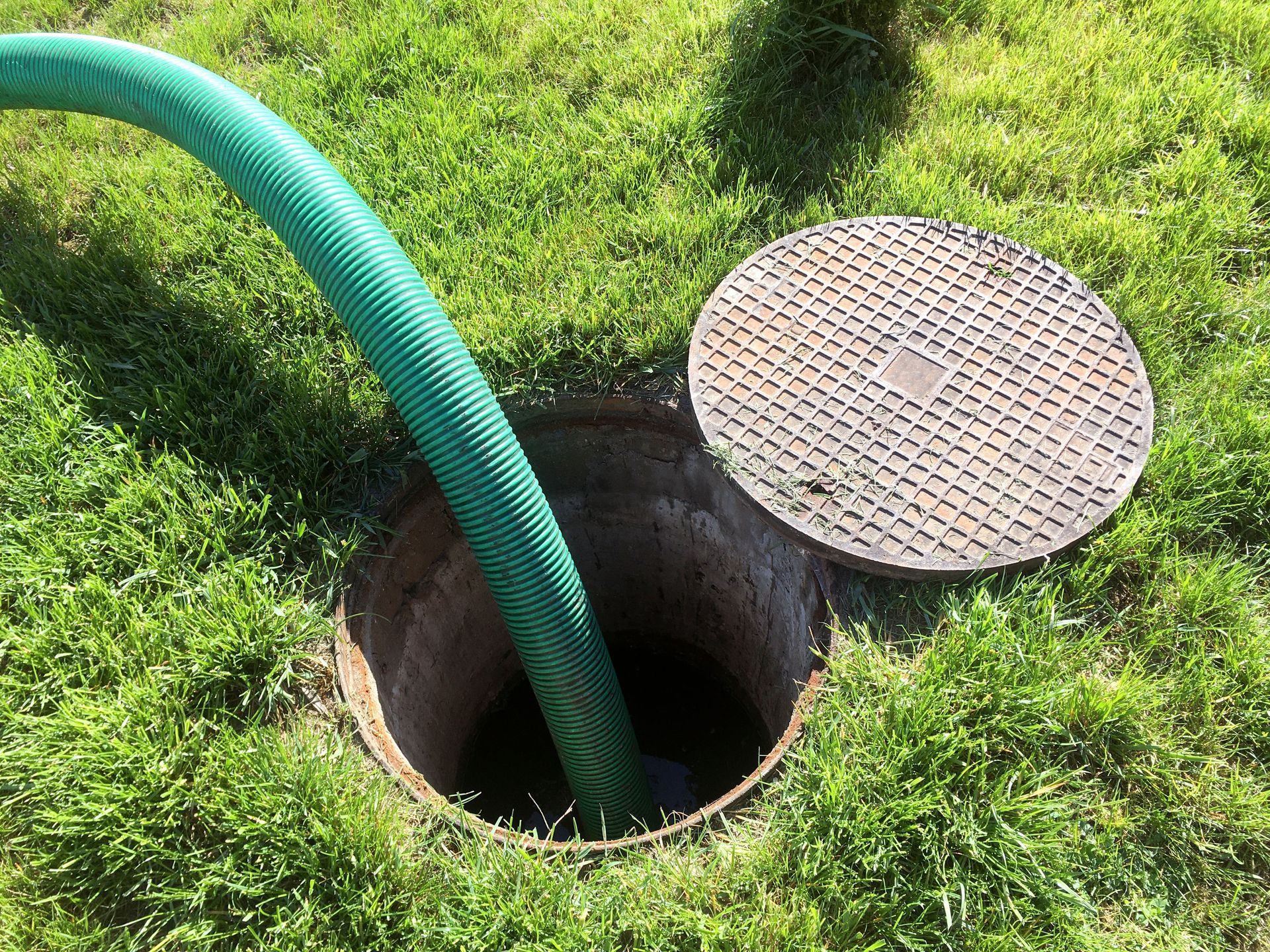 Green hose in an open septic tank on a grassy lawn; lid nearby.