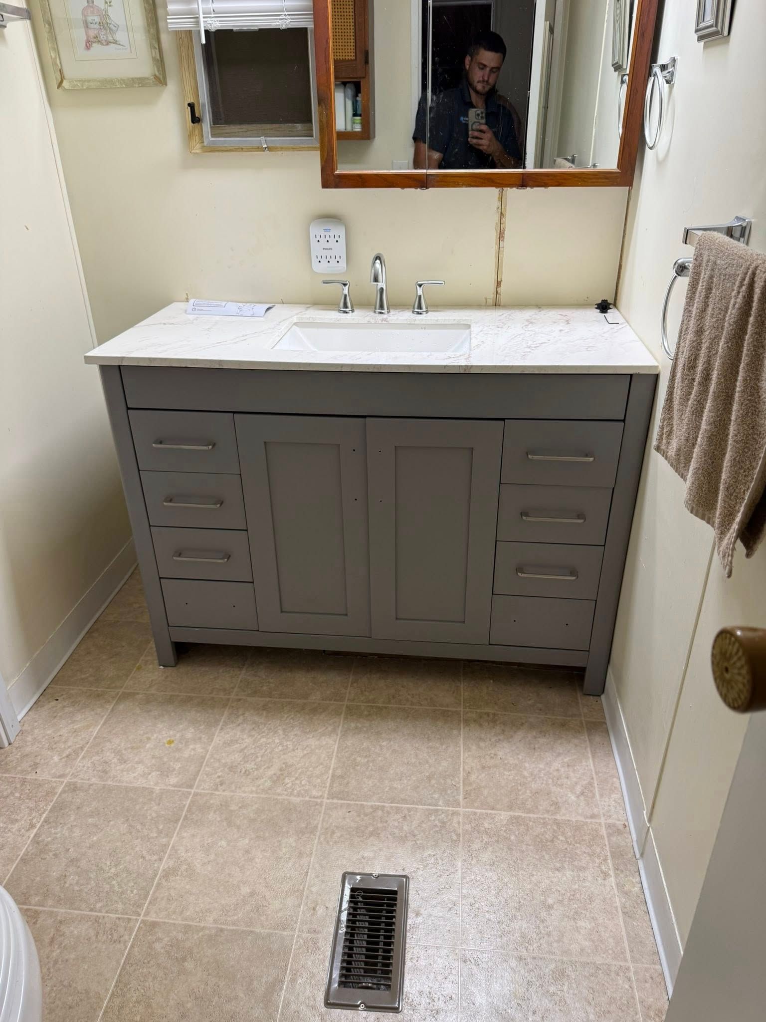 Gray bathroom vanity with white countertop, brown tile floor, and mirror reflecting a person.