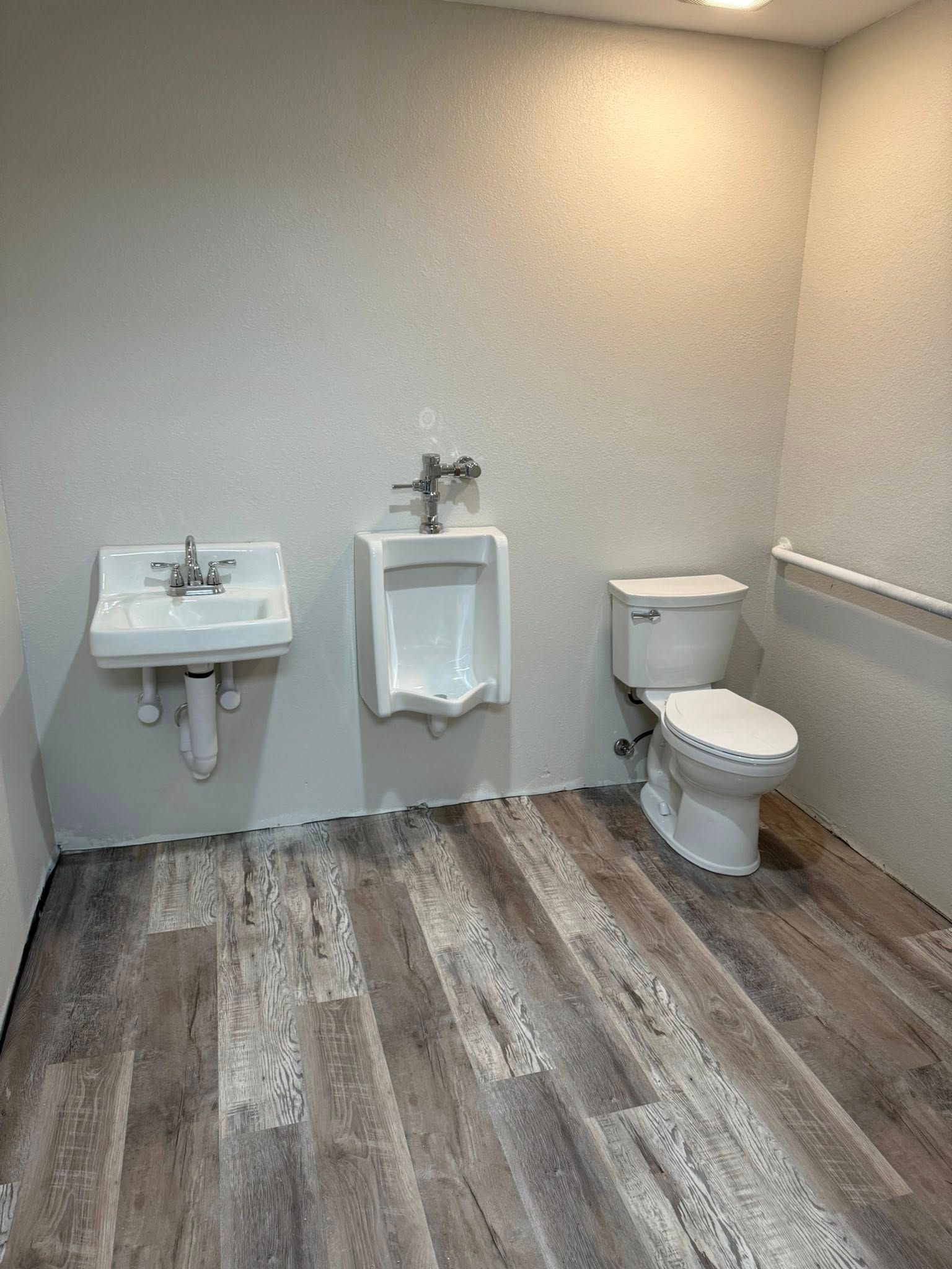 A small public restroom with a sink, urinal, and toilet on wood-look flooring. White walls and fixtures.
