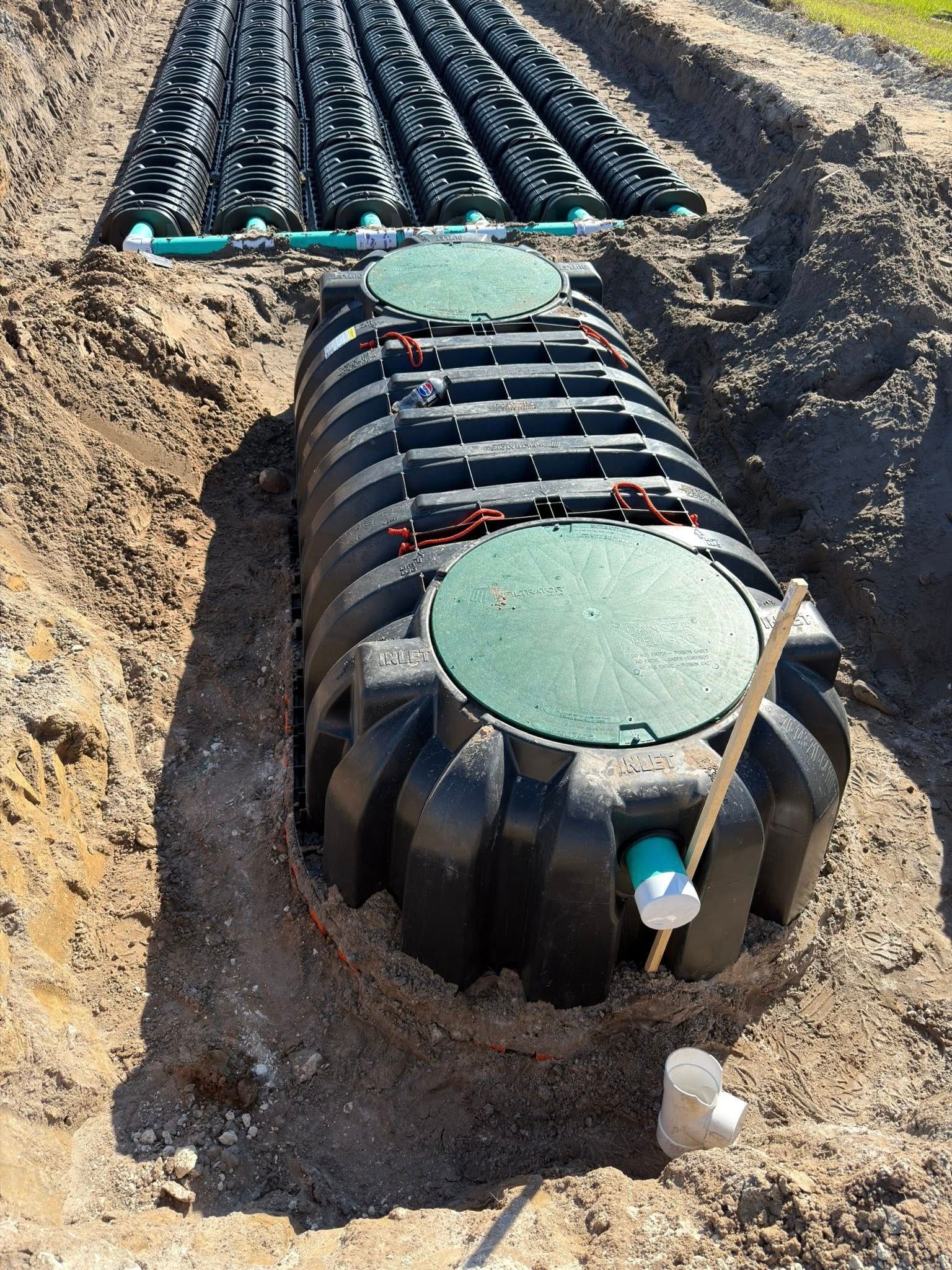 Septic system installation: black tank with green lids and connected pipes in an excavated trench.
