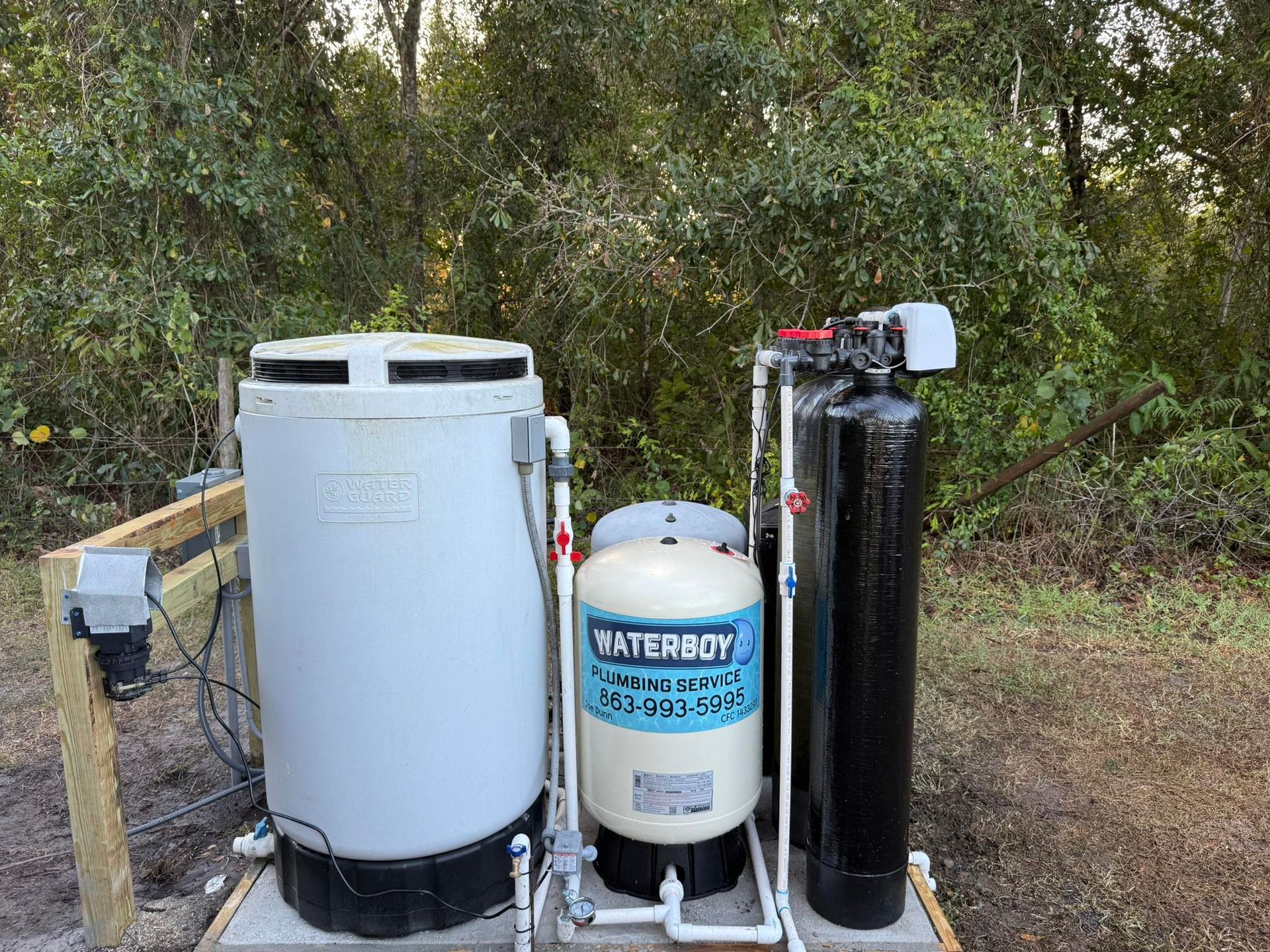 Water filtration system on a concrete base outdoors, with various tanks, pipes, and control box against a wooded backdrop.