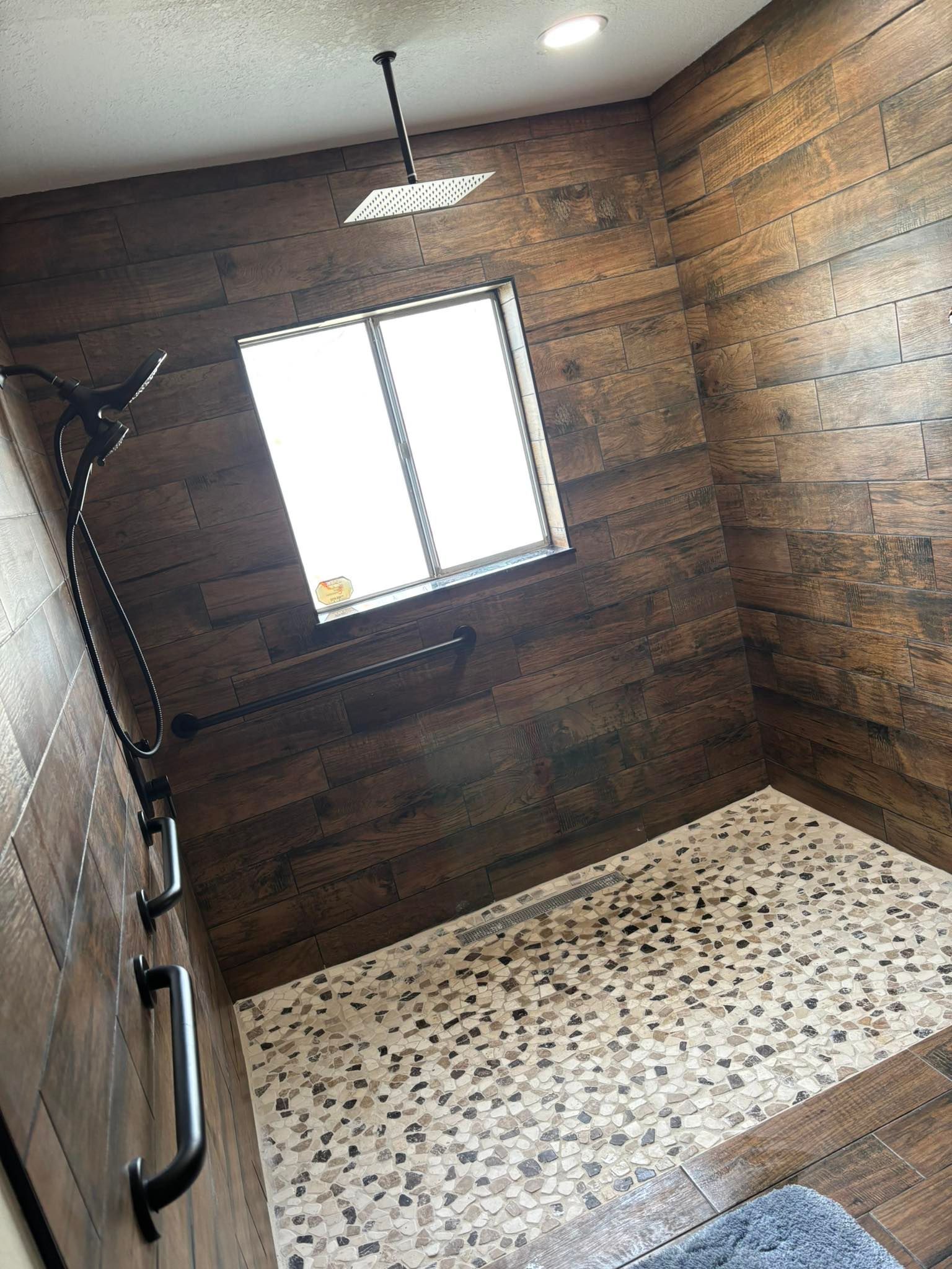 Bathroom with wood-look tile walls, pebble floor, and black shower fixtures. A window provides natural light.