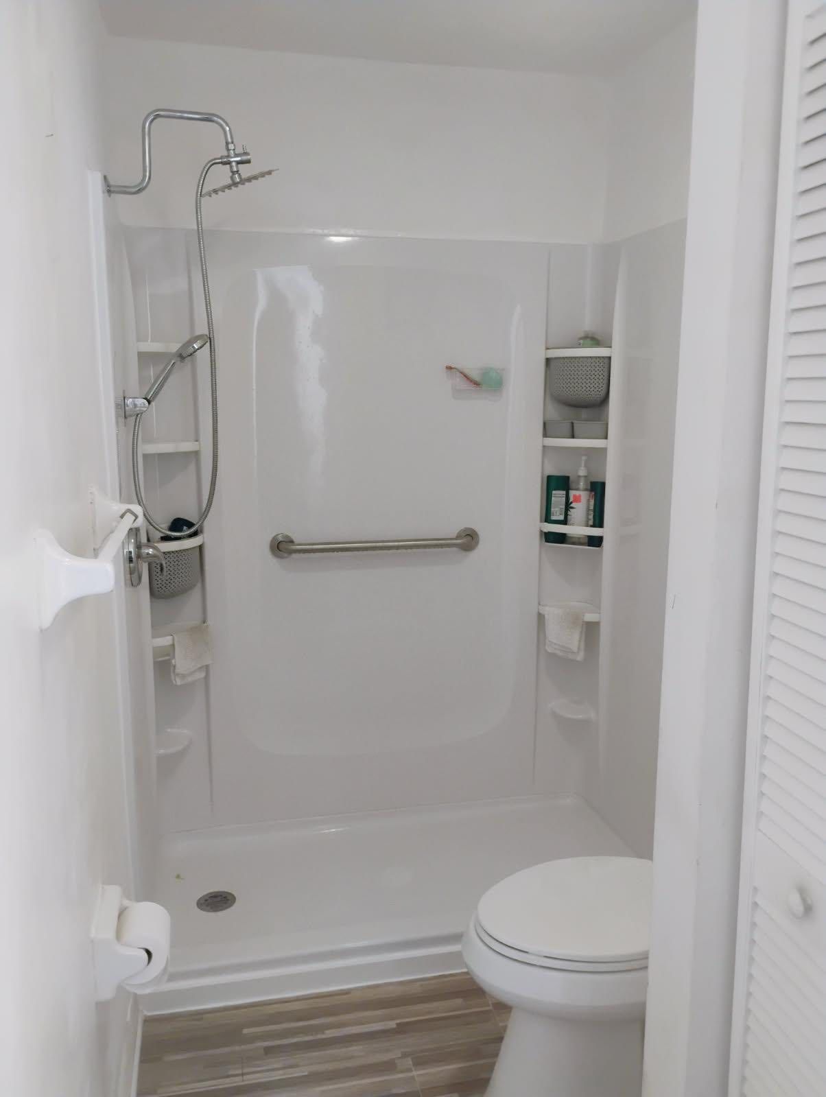 White bathroom with a walk-in shower, toilet, and shelving. Includes a grab bar.