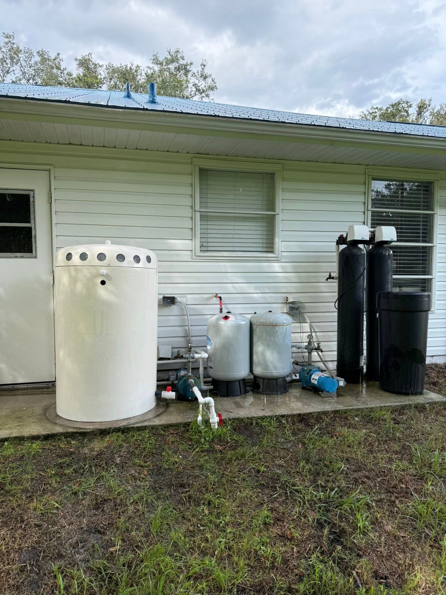 Outdoor water treatment system with several tanks, pipes, and pumps near a house.