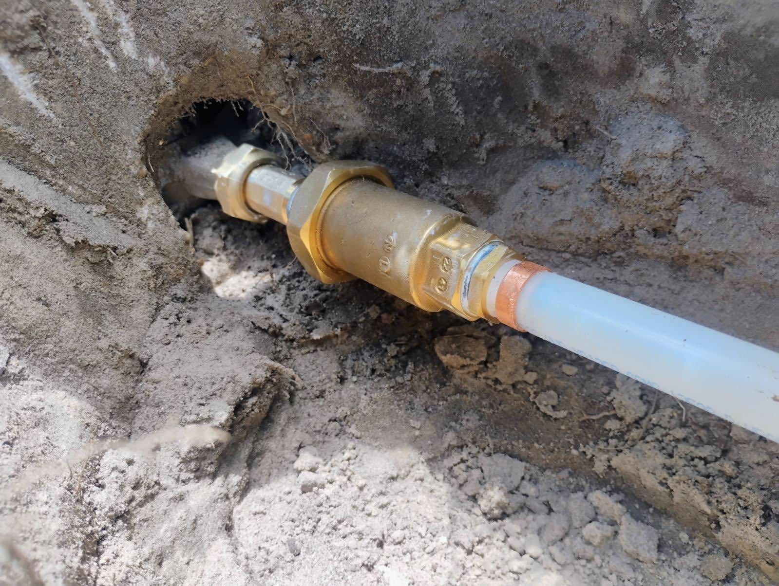 Brass plumbing fitting connecting white PEX pipe in a dirt trench.