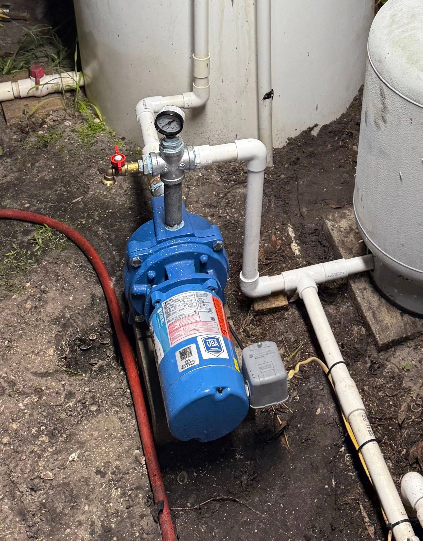 Blue water pump system with white pipes, gauge, and red valve near two white tanks.