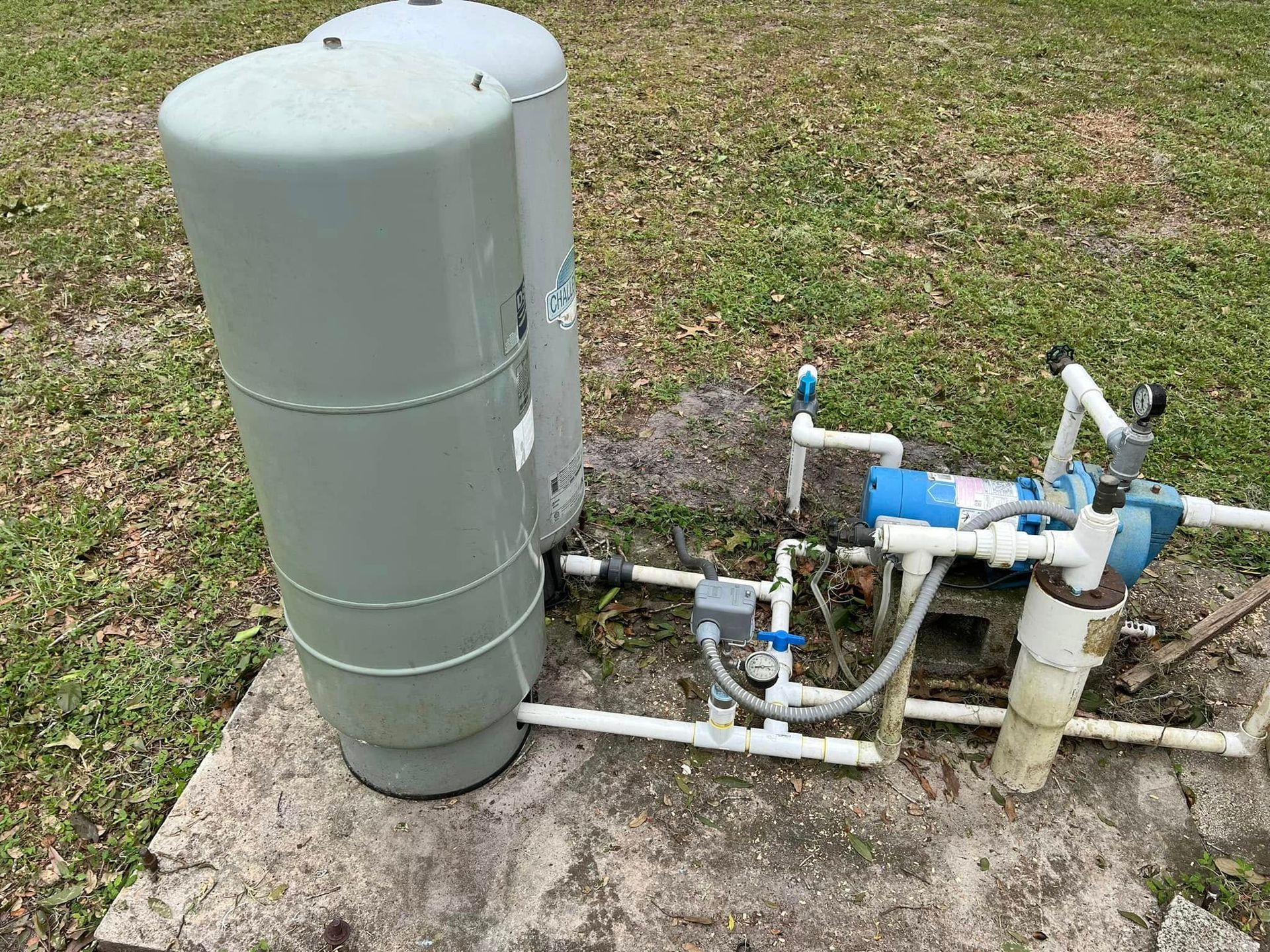 Well water system with two gray tanks, blue pump, white pipes, and concrete base outdoors.
