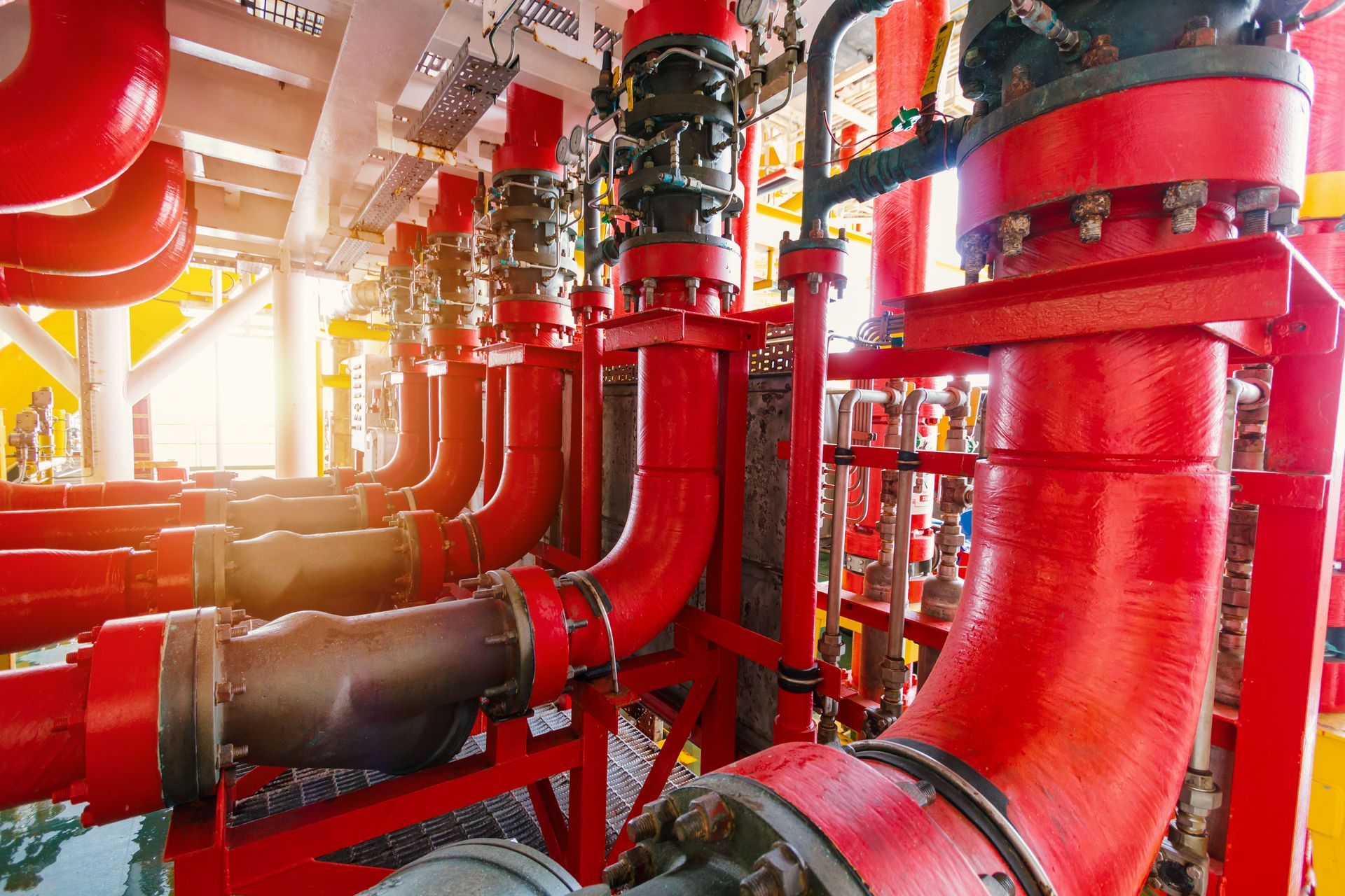 Bright red industrial pipes and valves in a mechanical plant interior, with sunlight streaming in from the left