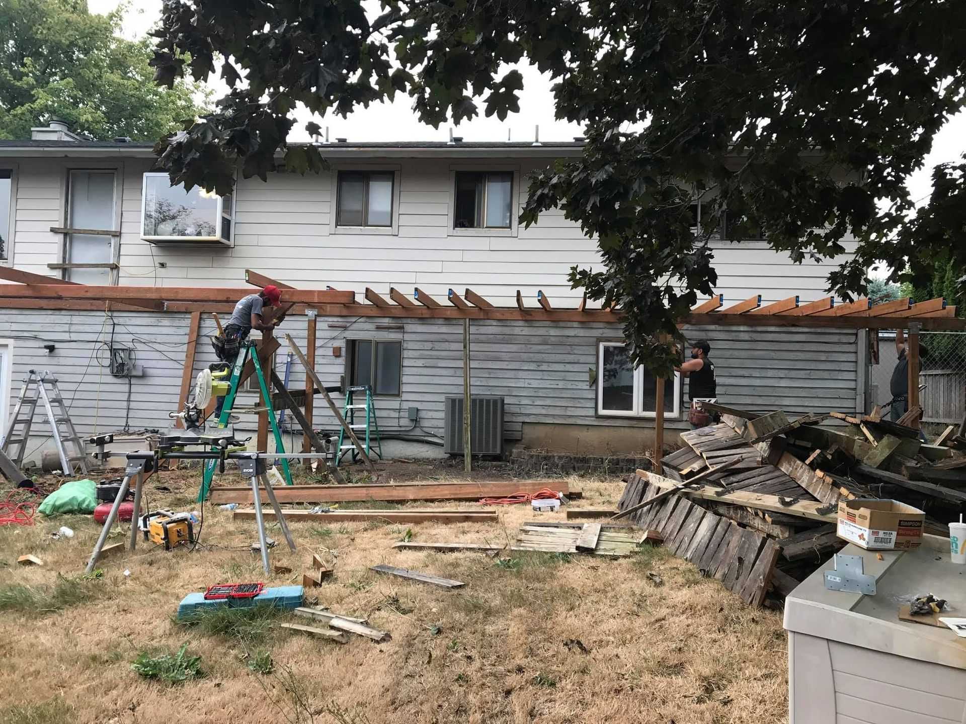 Backyard construction with ladder, tools, lumber, and a partially built pergola along a house wall