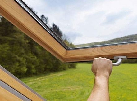 Hand opening a wooden skylight window above a green field and trees