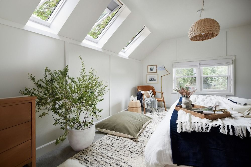 Bright attic bedroom with skylights, white walls, plants, and a neatly made bed by a window