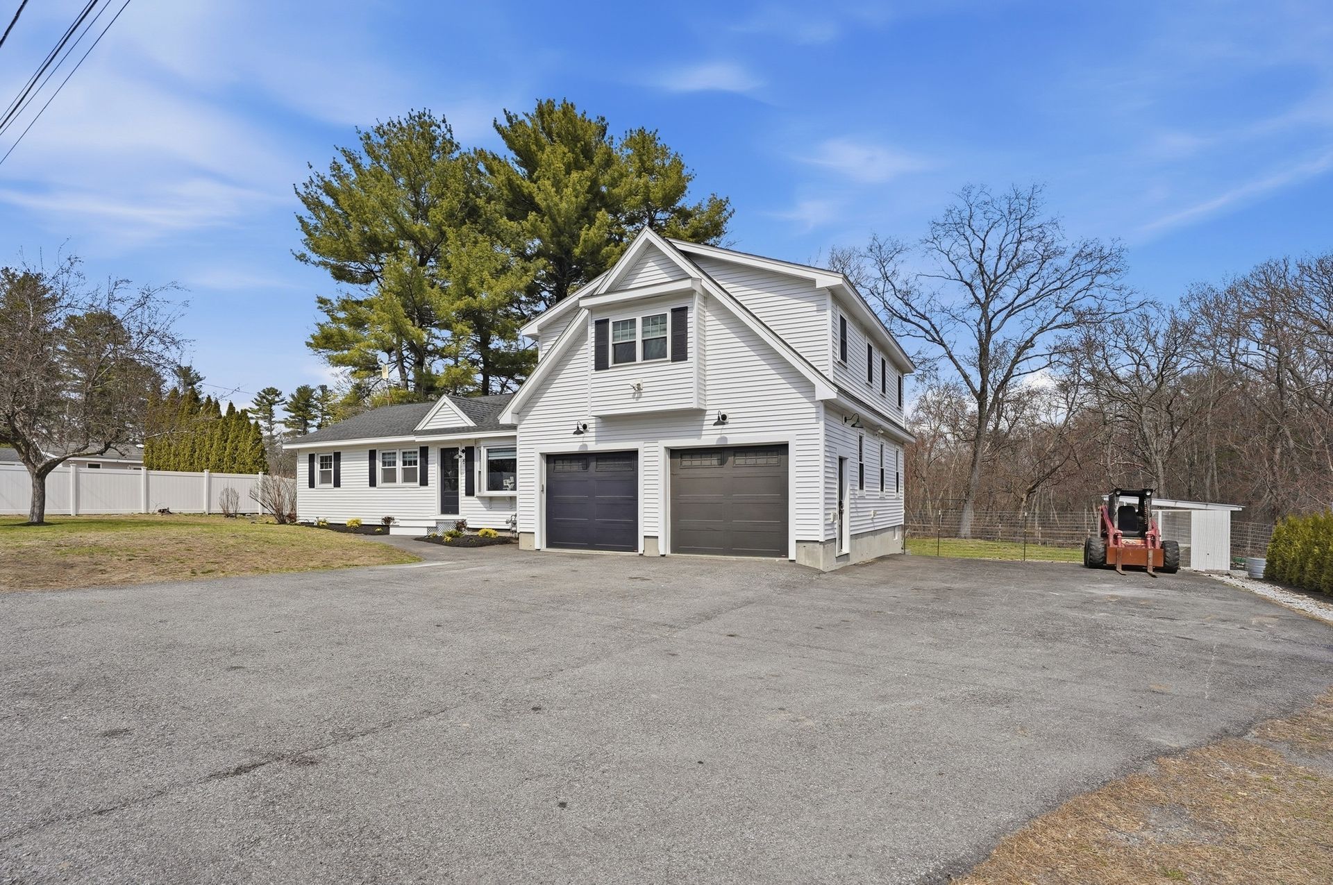 White house with two garage doors on a gravel lot, trees and a small tractor in the background.