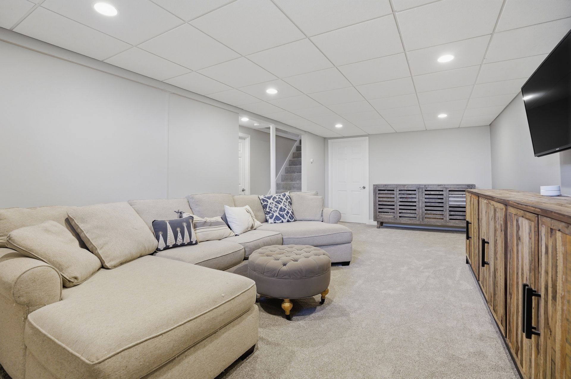 Bright basement living room with beige sectional, ottoman, gray carpet, and wood TV console