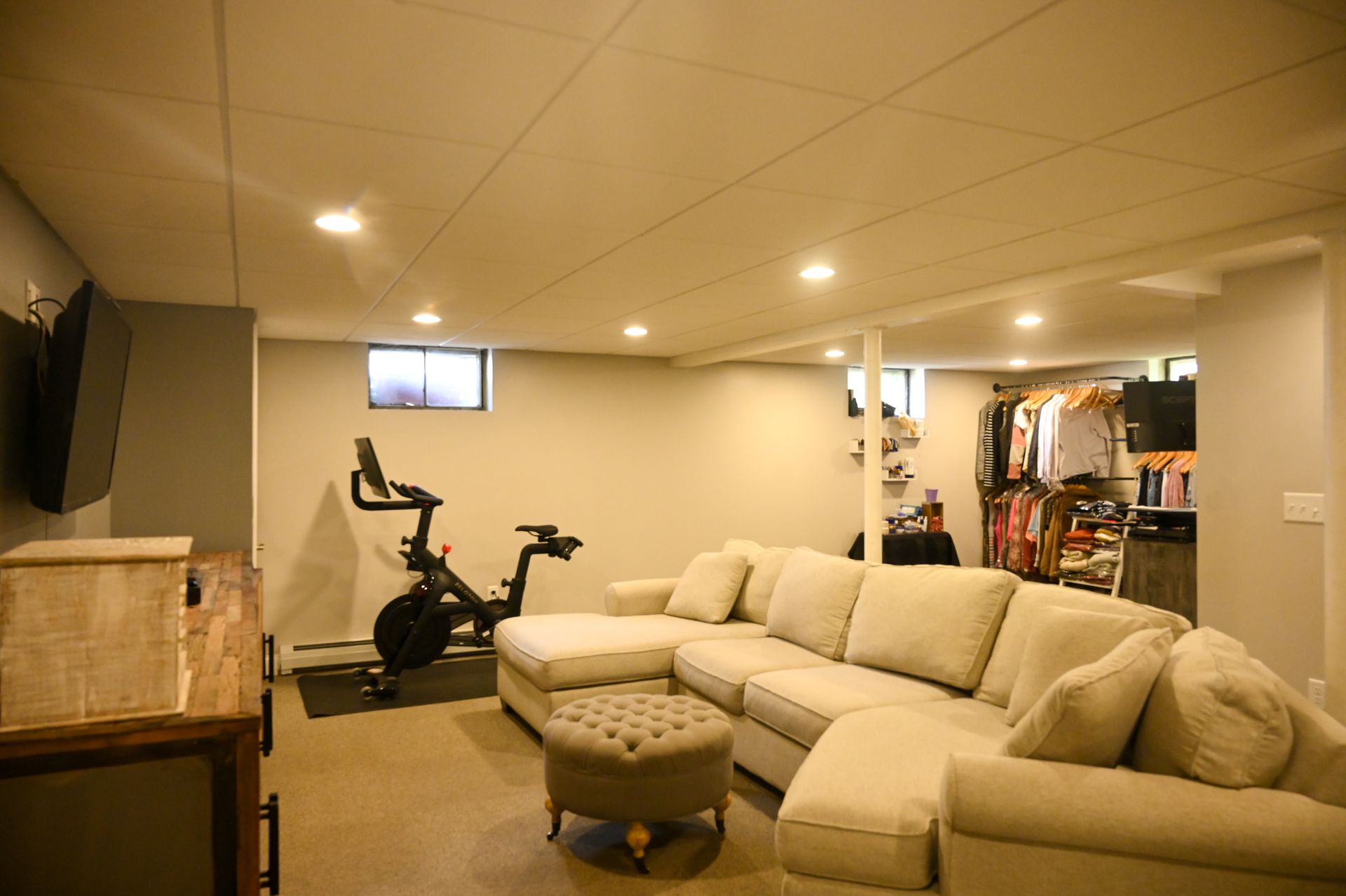 A carpeted basement with a beige sectional sofa, a tufted ottoman, a stationary exercise bike, and an open closet area.
