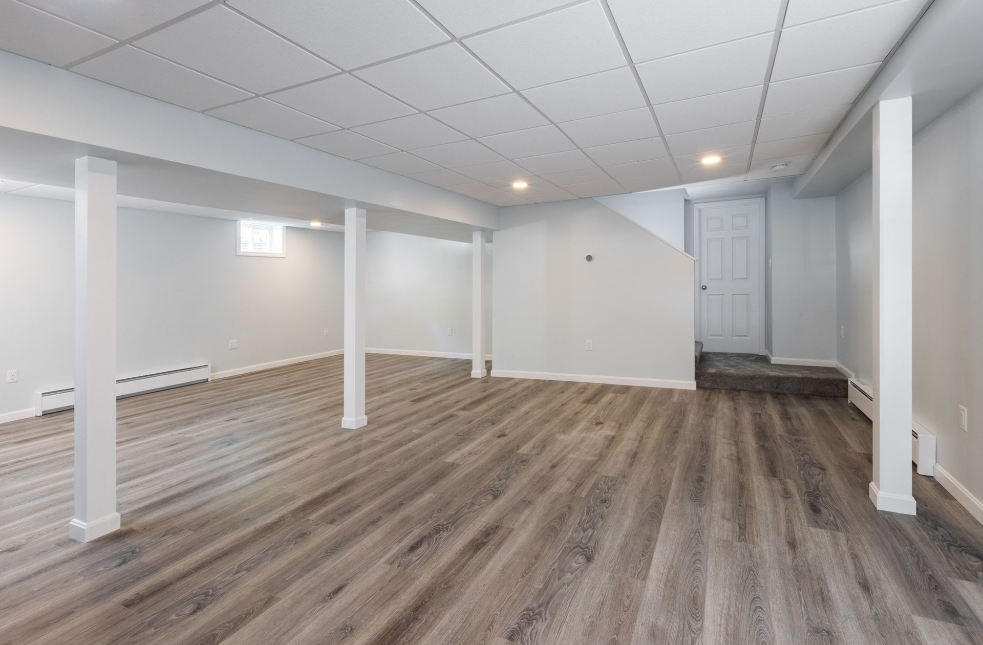 A basement interior featuring light gray walls, wood-look flooring, a drop ceiling, and white structural support columns.