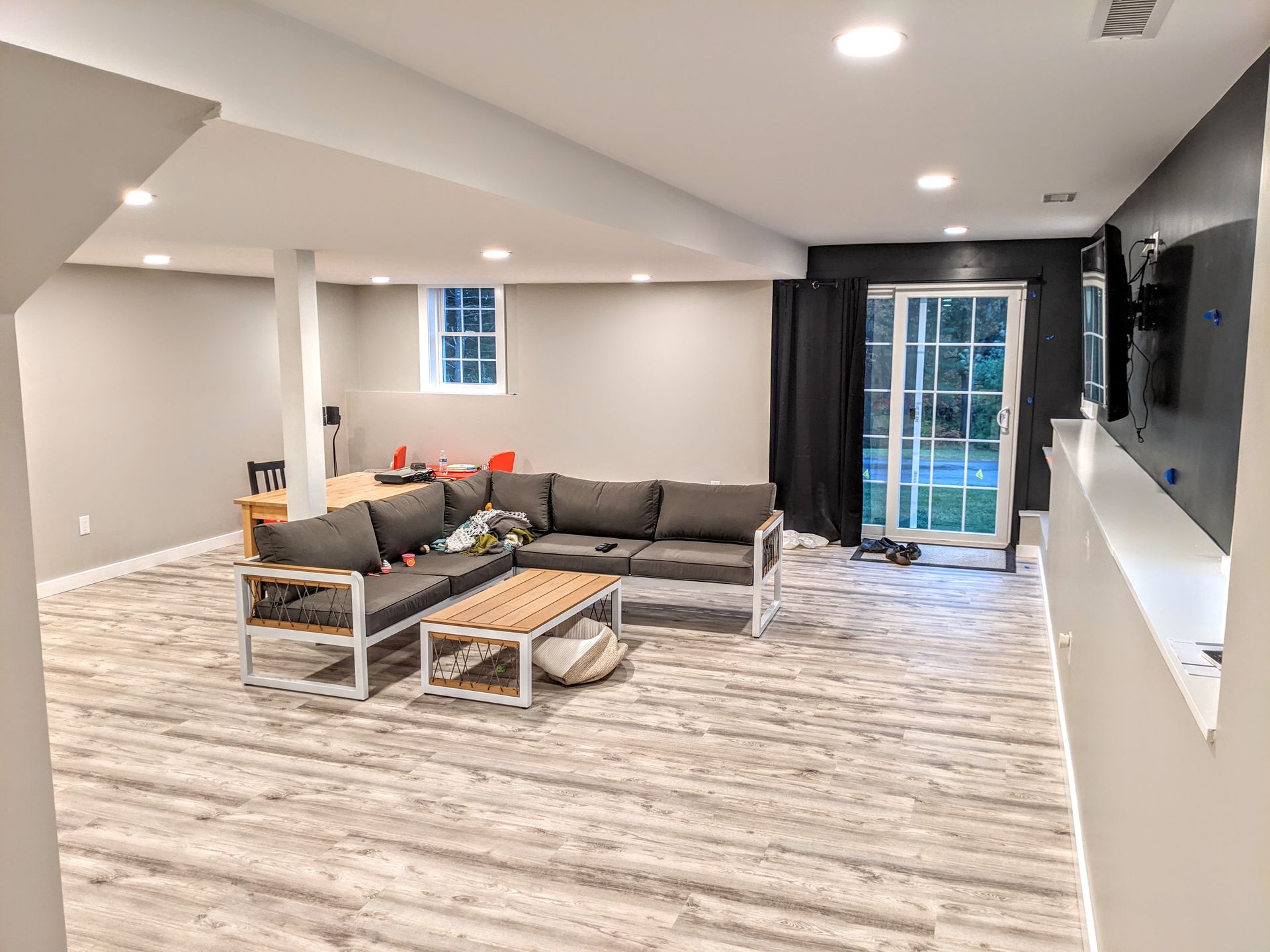 A modern basement with light gray wood-look flooring, a gray sectional sofa, a coffee table, and a glass sliding door.