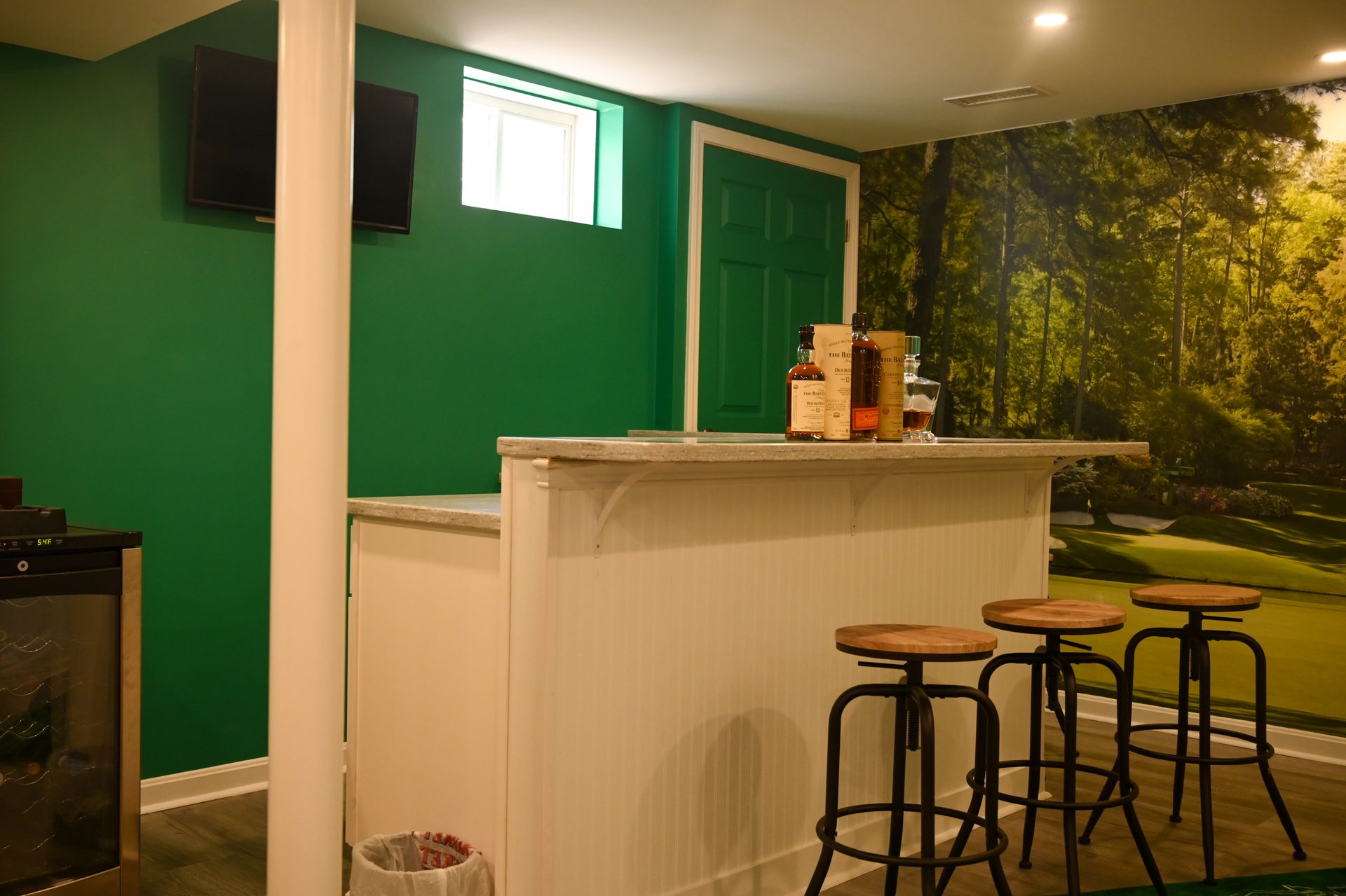 A white bar with three stools stands in front of a green wall and a forest mural in a basement room.
