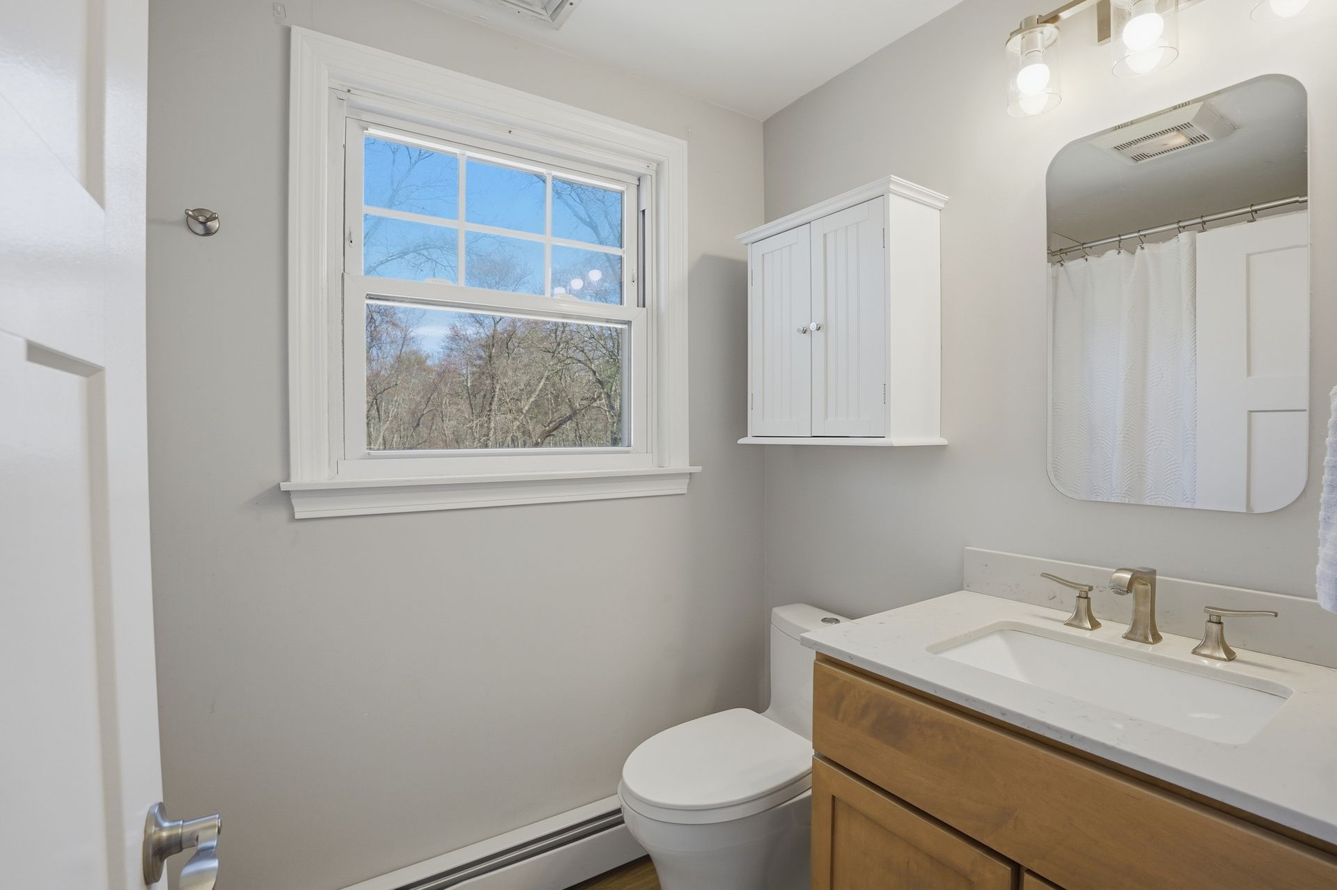 Small bathroom with a window, white walls, vanity, toilet, cabinet, and mirror lights.