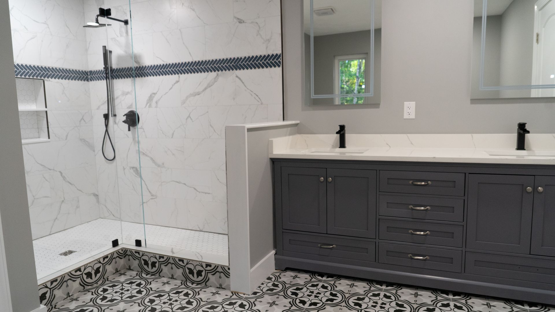 Modern bathroom with a tiled walk-in shower featuring patterned floor tiles and a gray double vanity with black faucets.