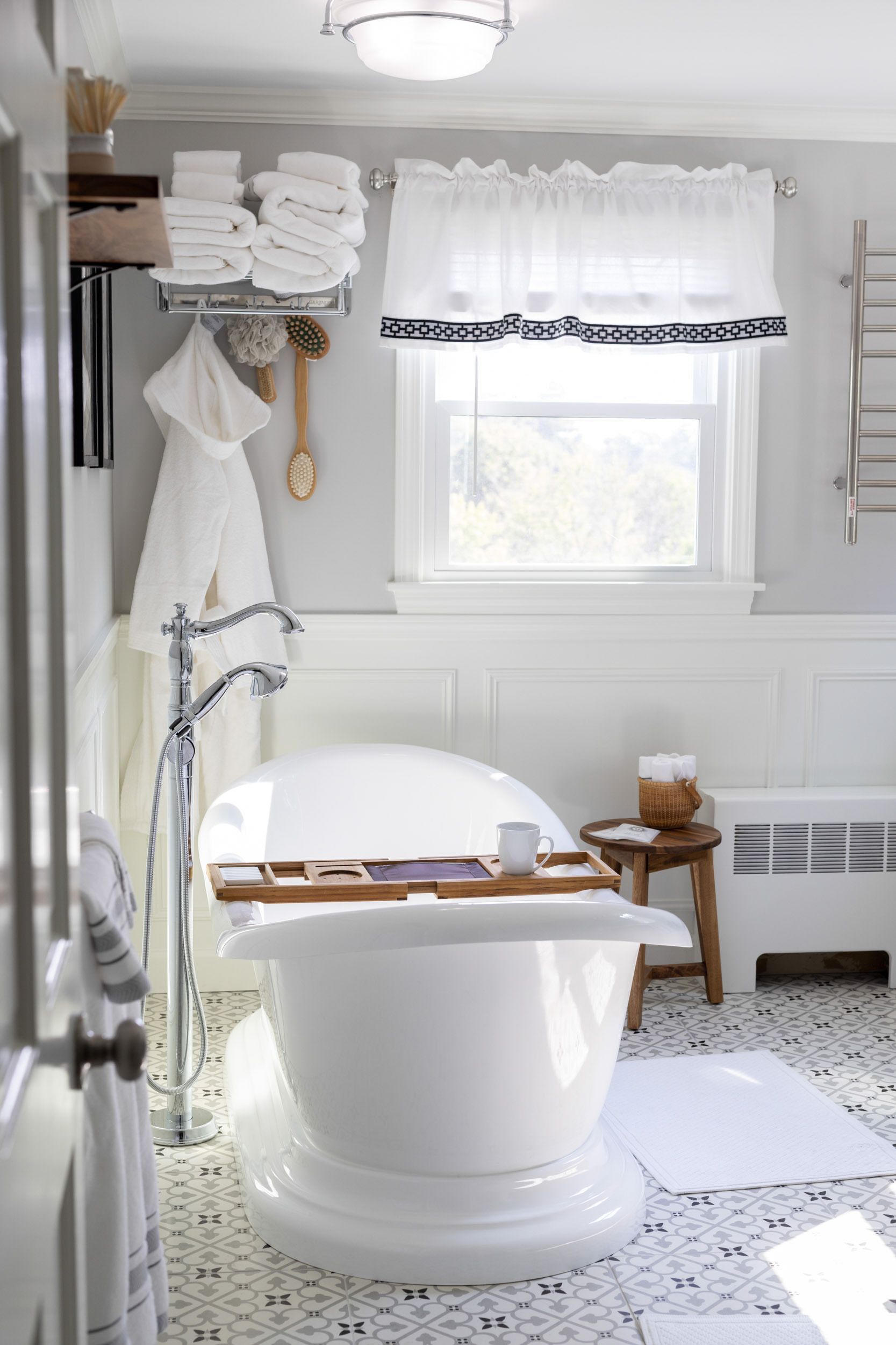 A white freestanding bathtub sits in a bright bathroom with a tiled floor, white wainscoting, and a window with a curtain.