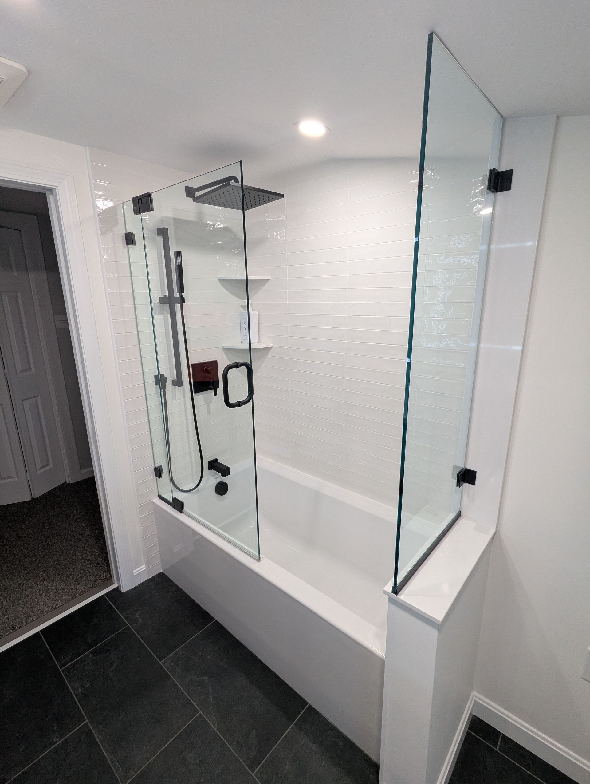 A modern bathroom features a white soaking tub with a glass panel enclosure, black hardware, and textured white tile walls.