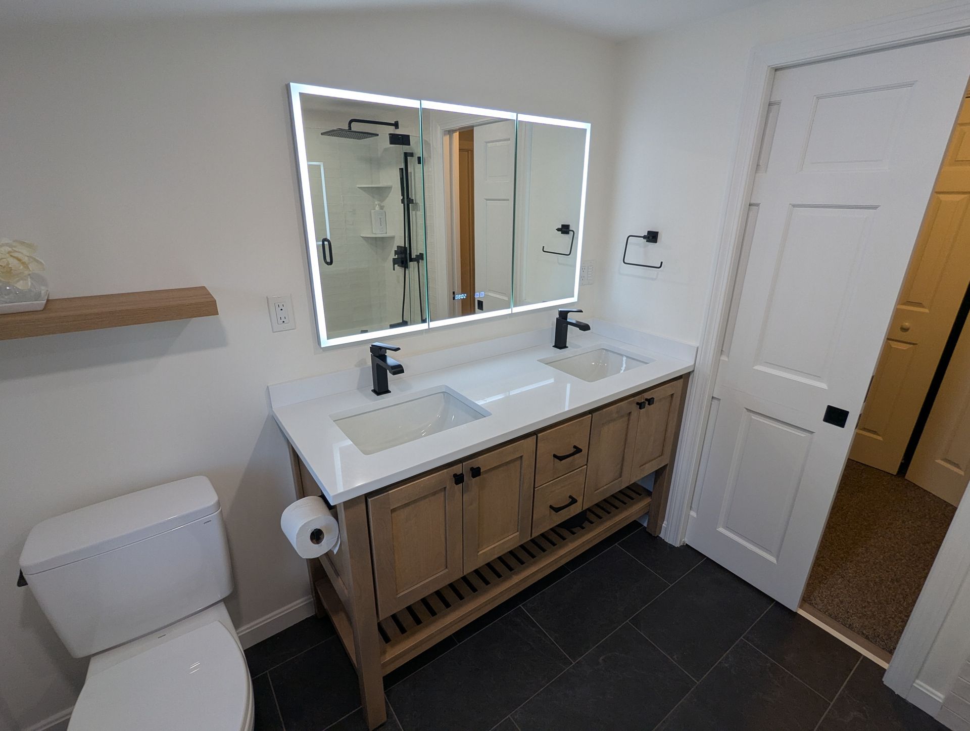 A modern bathroom with a double-sink wood vanity, an illuminated mirror, white walls, and dark tile flooring.