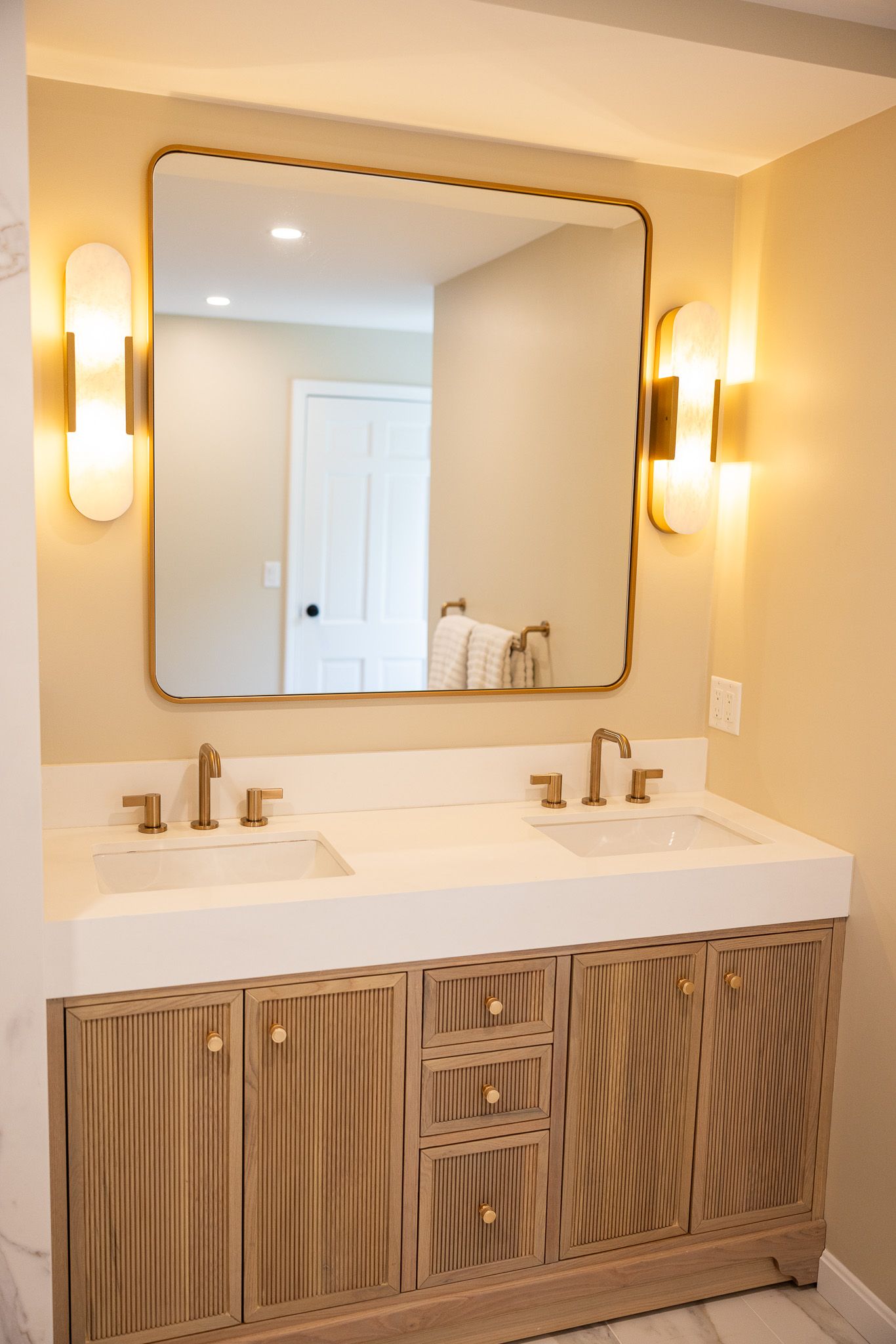 A bathroom vanity with a white double-sink countertop, a rectangular mirror, and two wall-mounted lights in a gold finish.