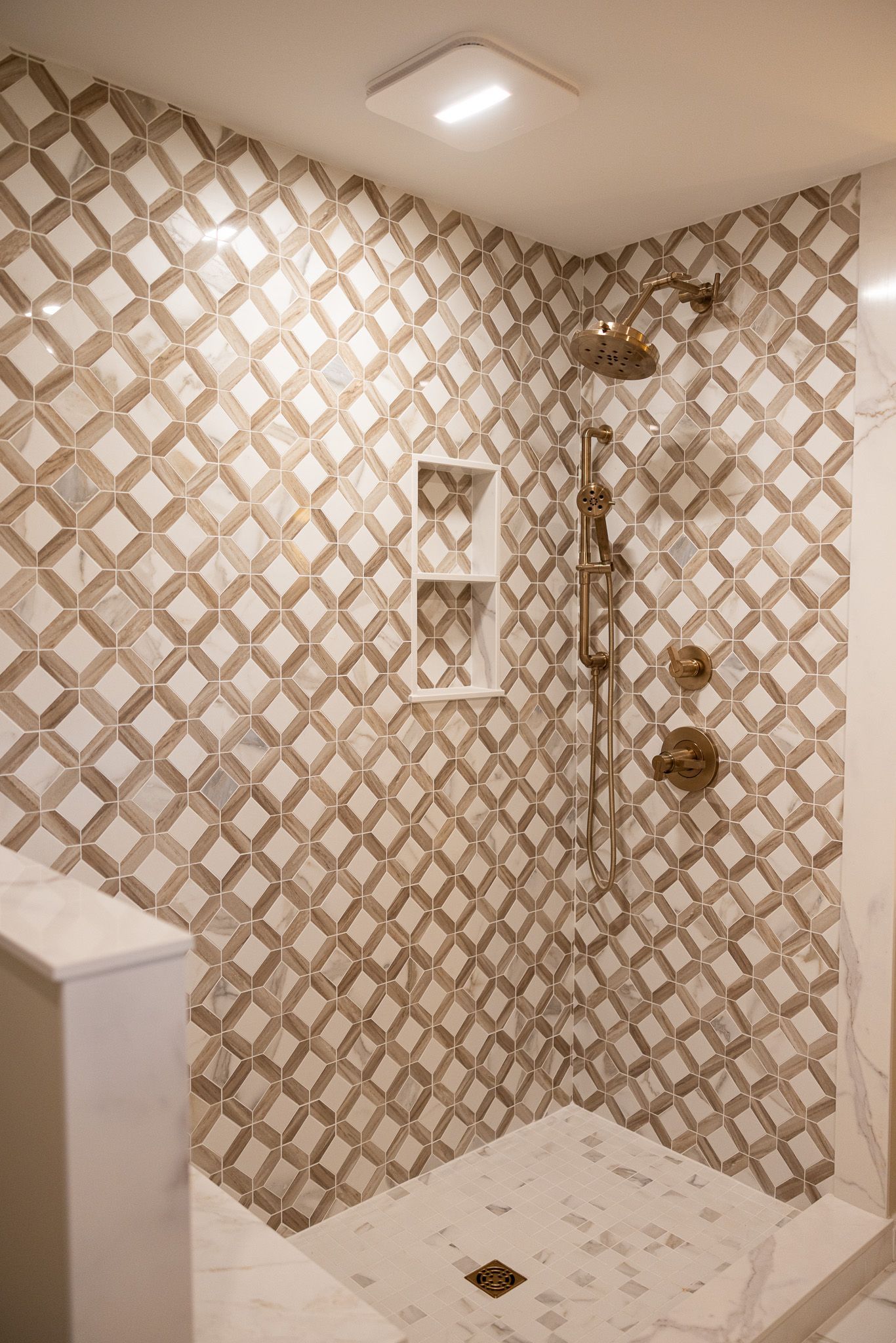 Shower stall with white and gold geometric patterned tile walls, a built-in shelf, and gold plumbing fixtures.