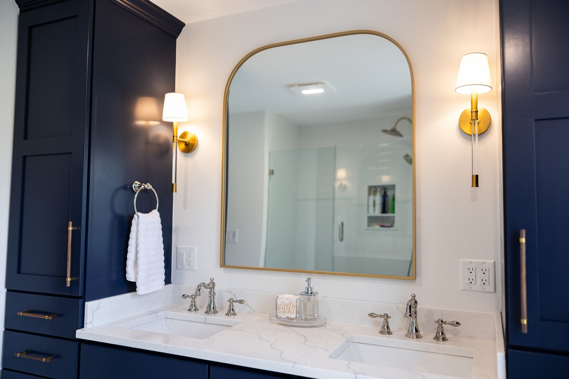 Double bathroom vanity with navy cabinets, marble countertop, gold-rimmed arch mirror, and gold wall sconces.
