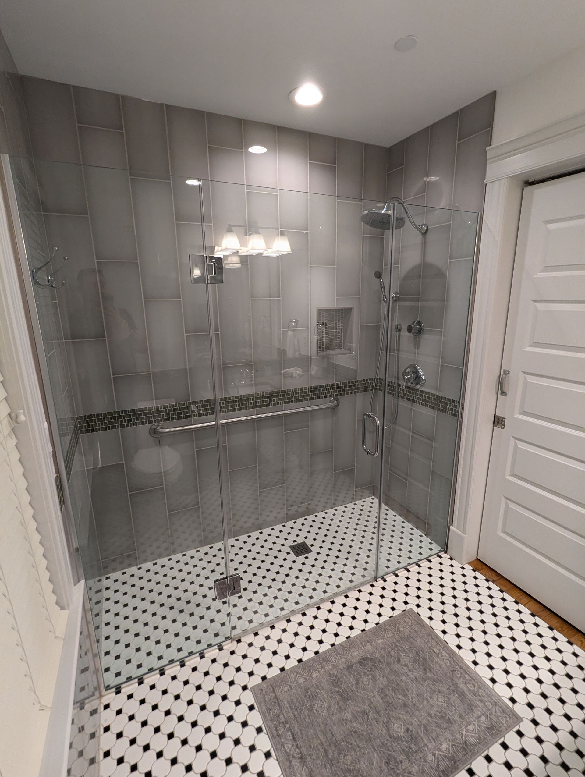 A modern walk-in shower with grey subway tile walls, a mosaic accent strip, glass doors, and a black-and-white floor.