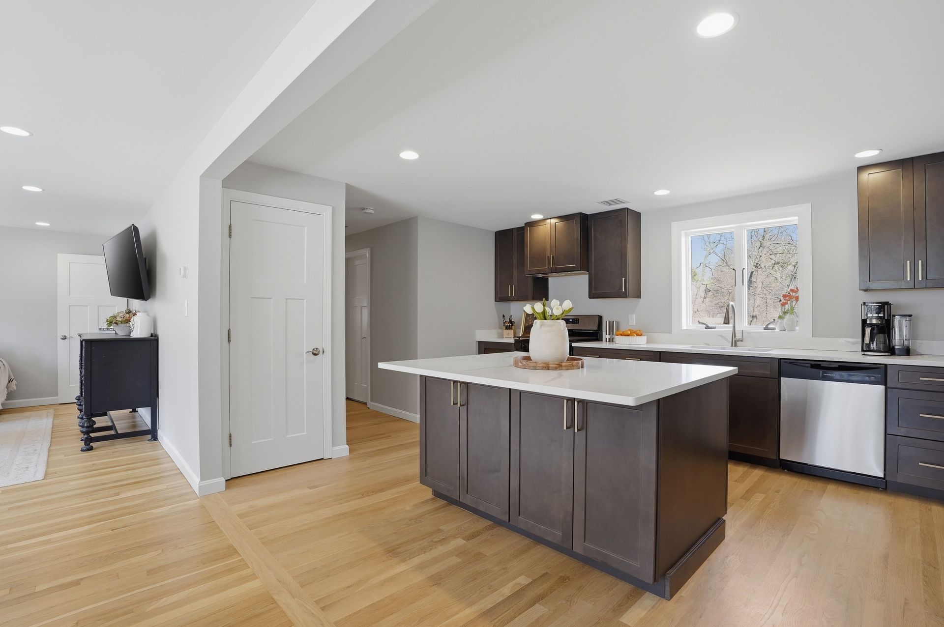 Bright modern kitchen with dark cabinets, white island, wood floors, and adjacent living area