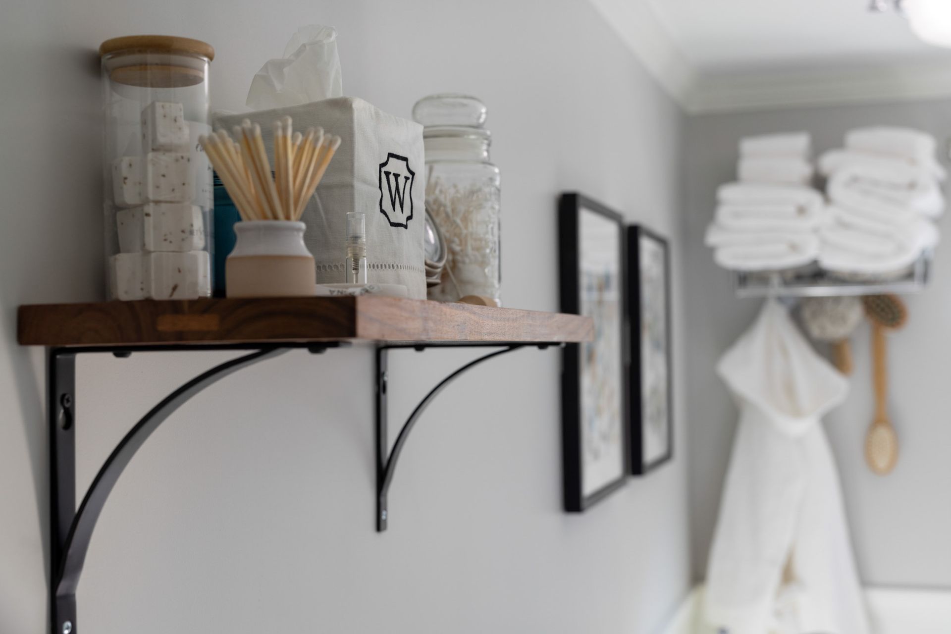 A bathroom wall with a wooden shelf holding glass jars and toiletries, next to framed art and stacked towels on a rack.