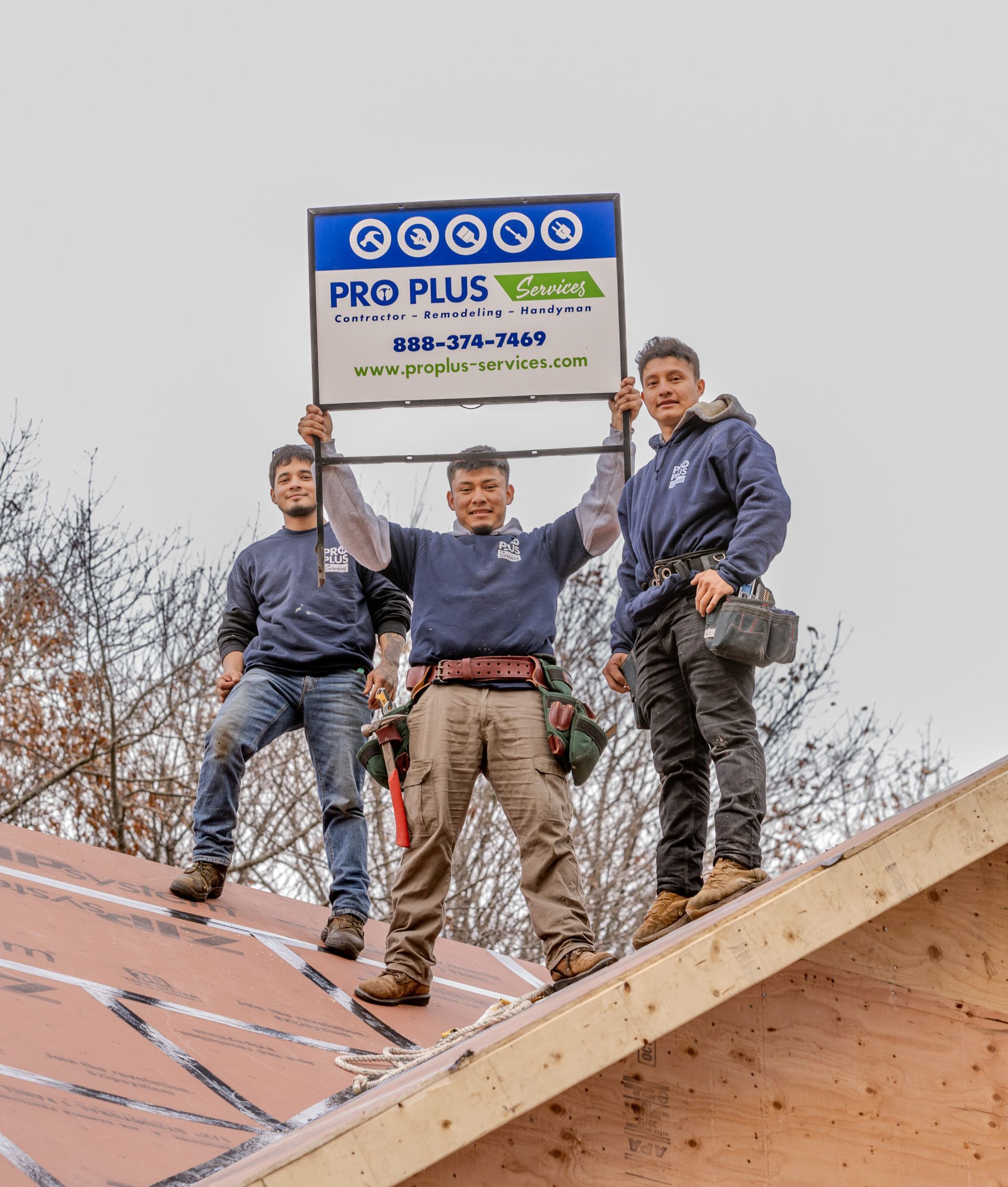 Three workers stand on a roof framing structure, smiling as they hold up a Pro Plus Construction company sign.