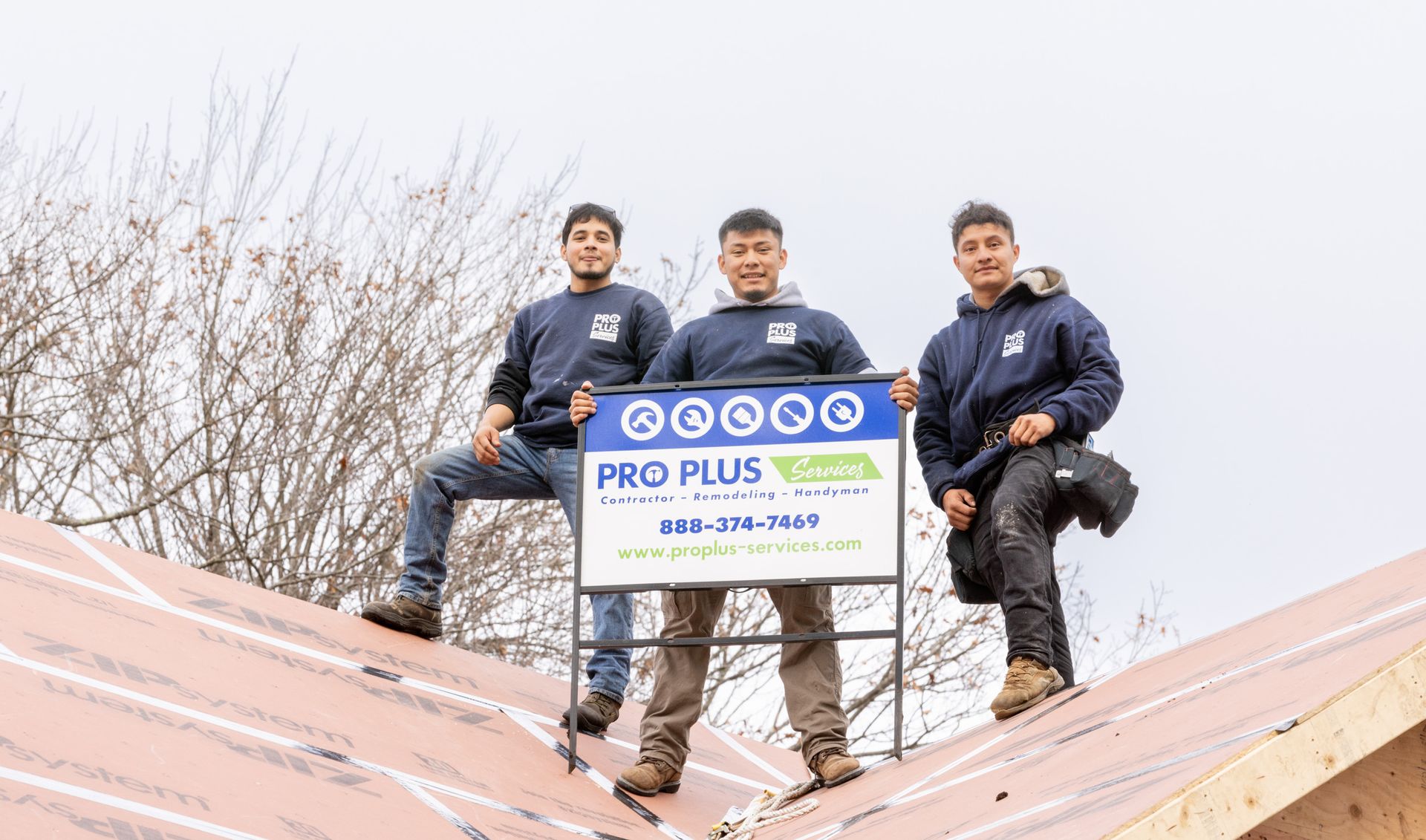 Three people holding a Pro Plus roofing company sign while standing on a roof under construction.