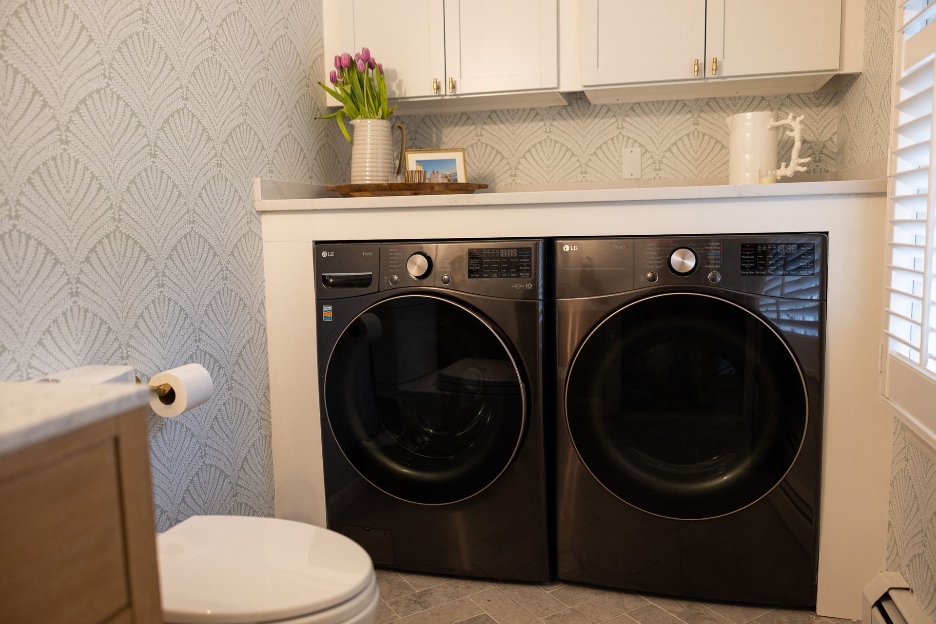 A laundry room area with dark front-load machines, white cabinetry above, and light blue patterned wallpaper.