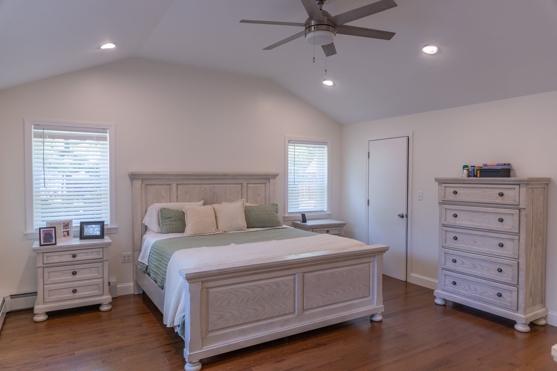 A bedroom with a wooden bed set, white bedding, green accent pillows, and windows on a light-colored wood floor.