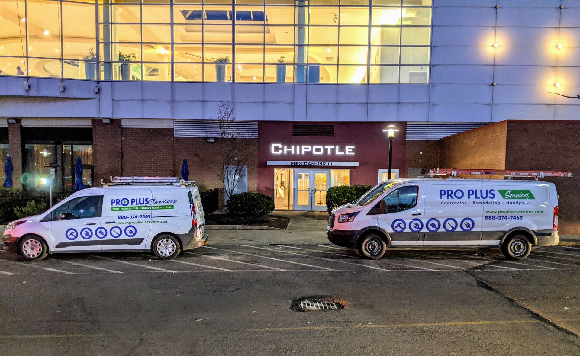 Two white Pro Plus Plumbing service vans parked in front of a Chipotle restaurant at dusk.