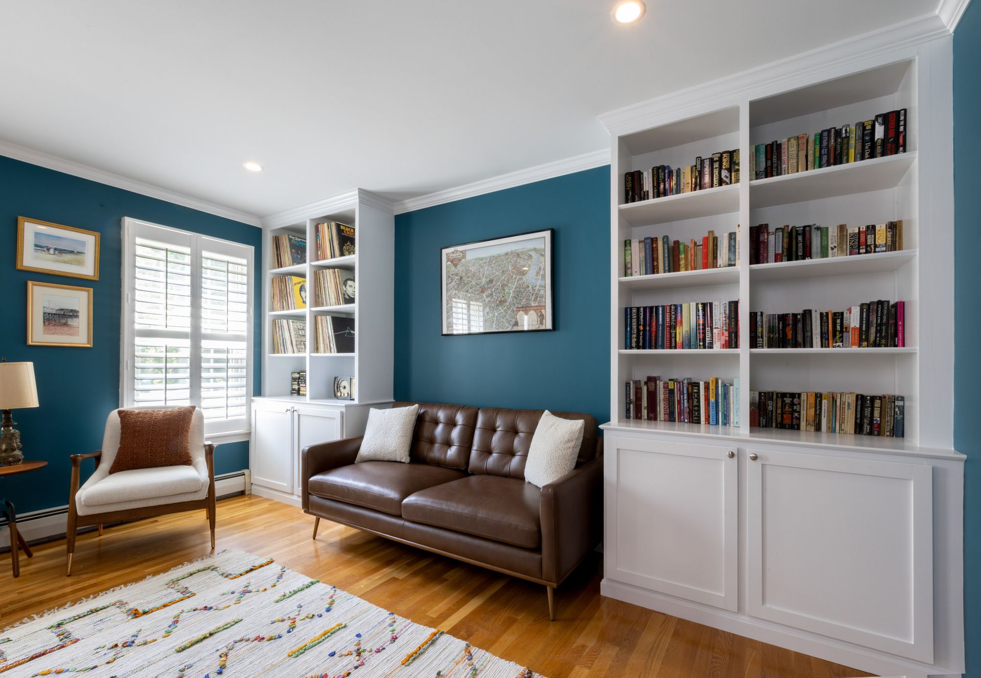 A cozy room with dark blue walls, built-in white bookshelves, a brown leather sofa, an accent chair, and a patterned rug.