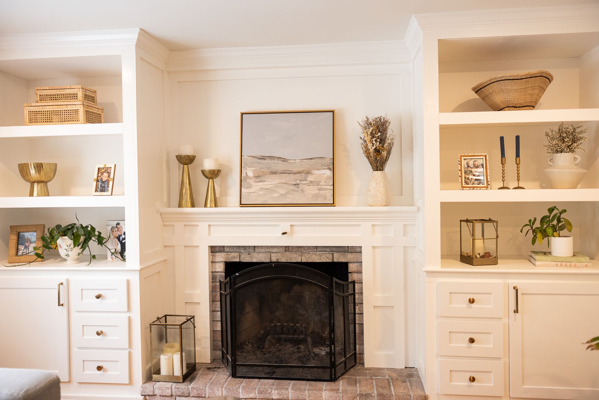 A fireplace mantel with white built-in shelves on both sides, decorated with vases, plants, candles, and framed art.