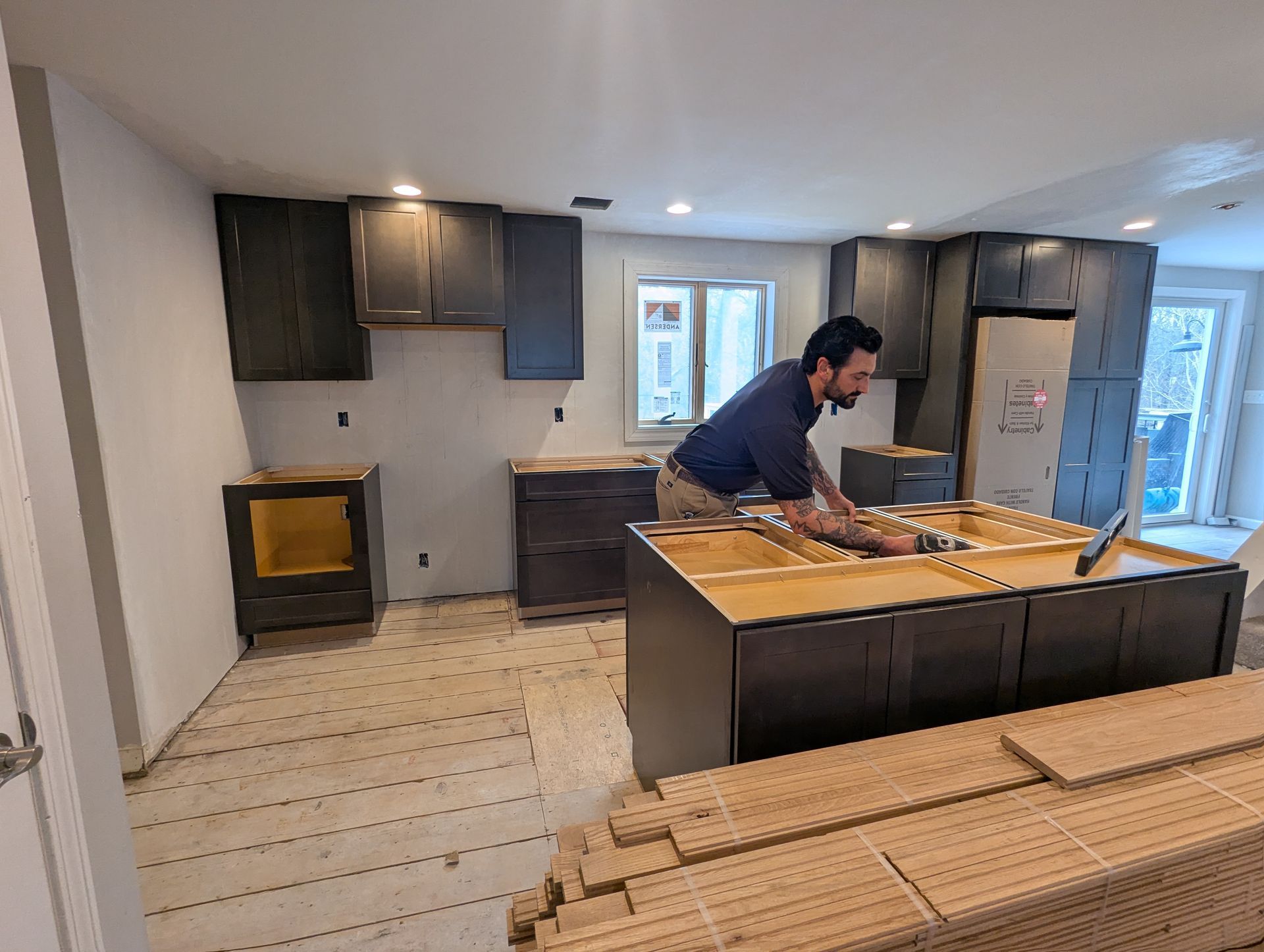 A person works on the kitchen island in a room featuring dark cabinets and unfinished flooring during a renovation.