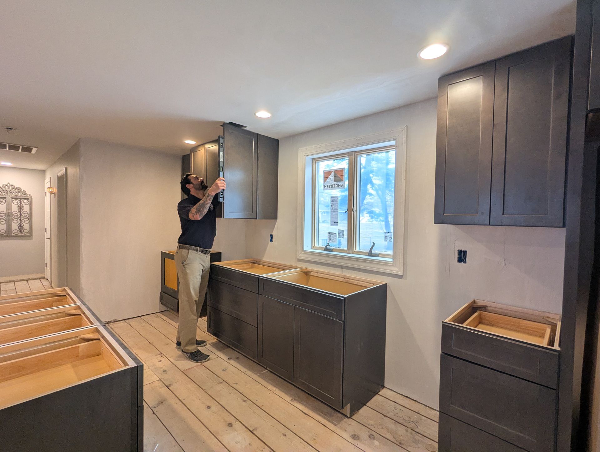 A person in a room under construction installs dark-colored cabinets on a wall with a window.