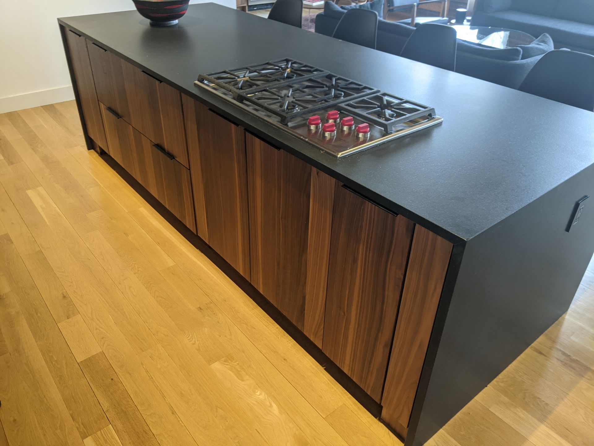 A dark, rectangular kitchen island with a built-in gas stove and warm-toned wood cabinetry, set on light wood flooring.