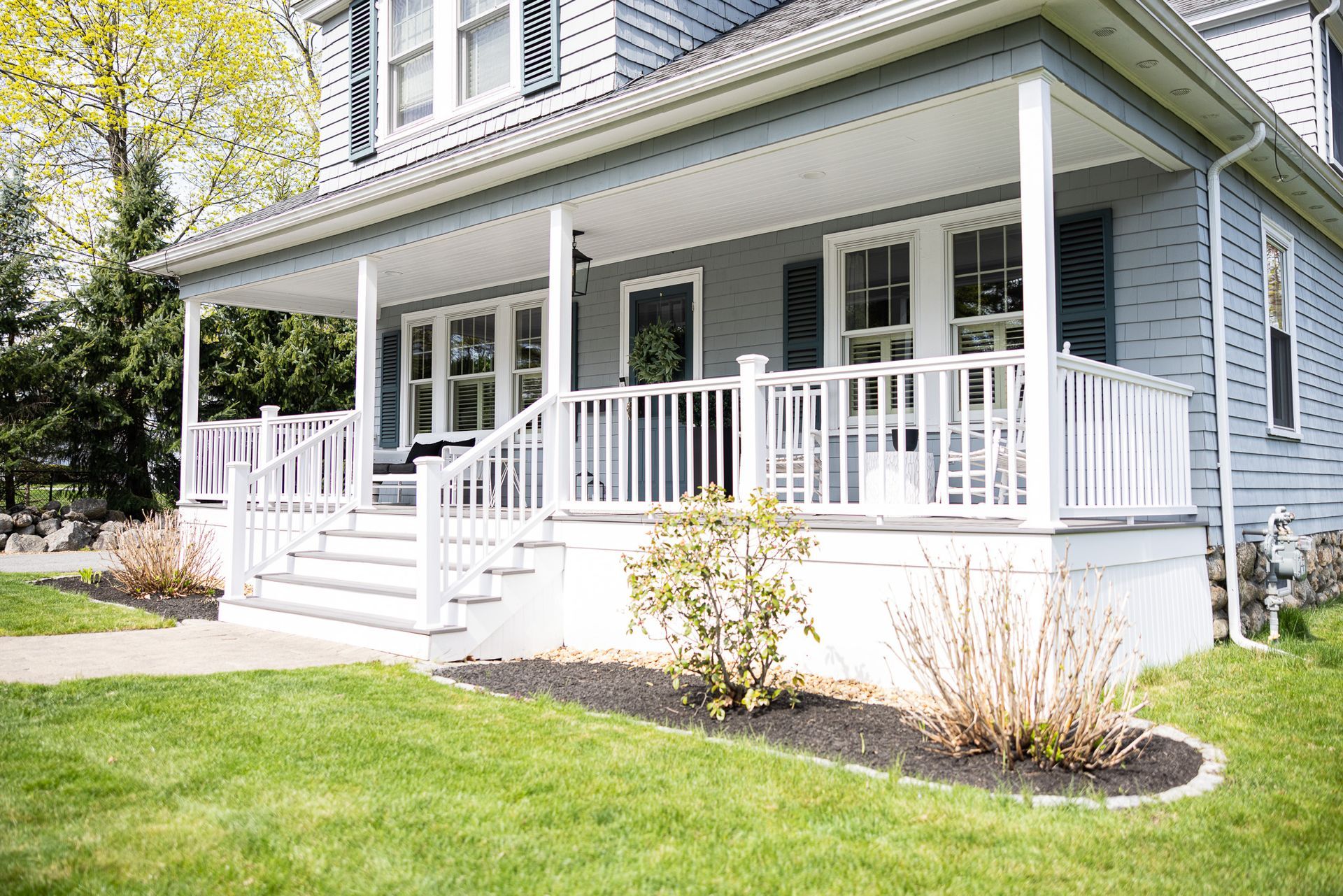 A gray house with a white wrap-around porch, white railing, stairs, and small landscaping bushes in the front yard.