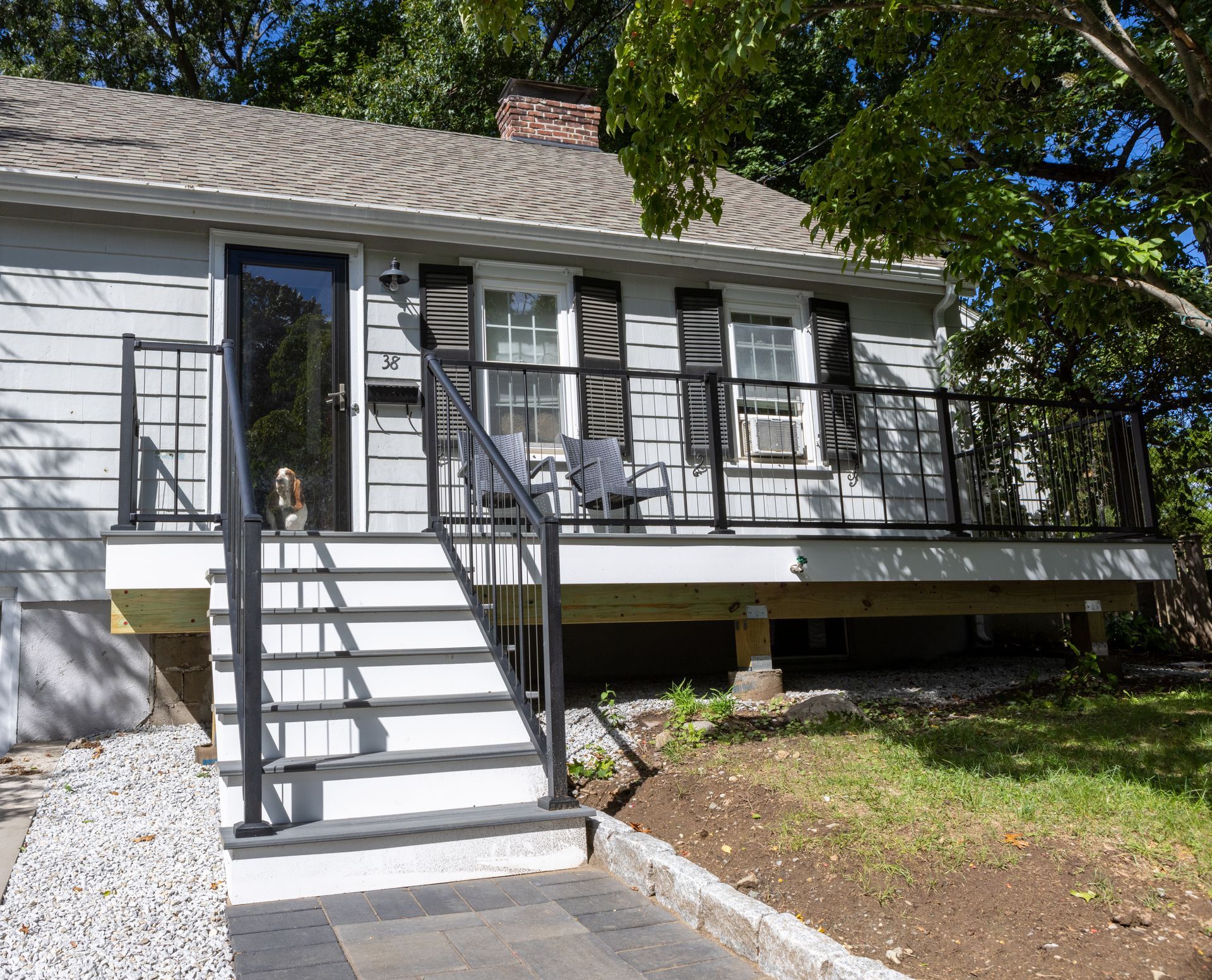 A gray house with a front deck, black railings, white stairs, and a dog standing by the front door.