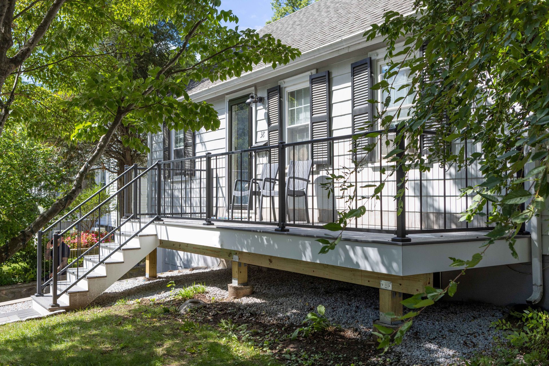 A gray cottage with a new wooden deck and black metal railing, accessed by a set of stairs from a gravel yard.