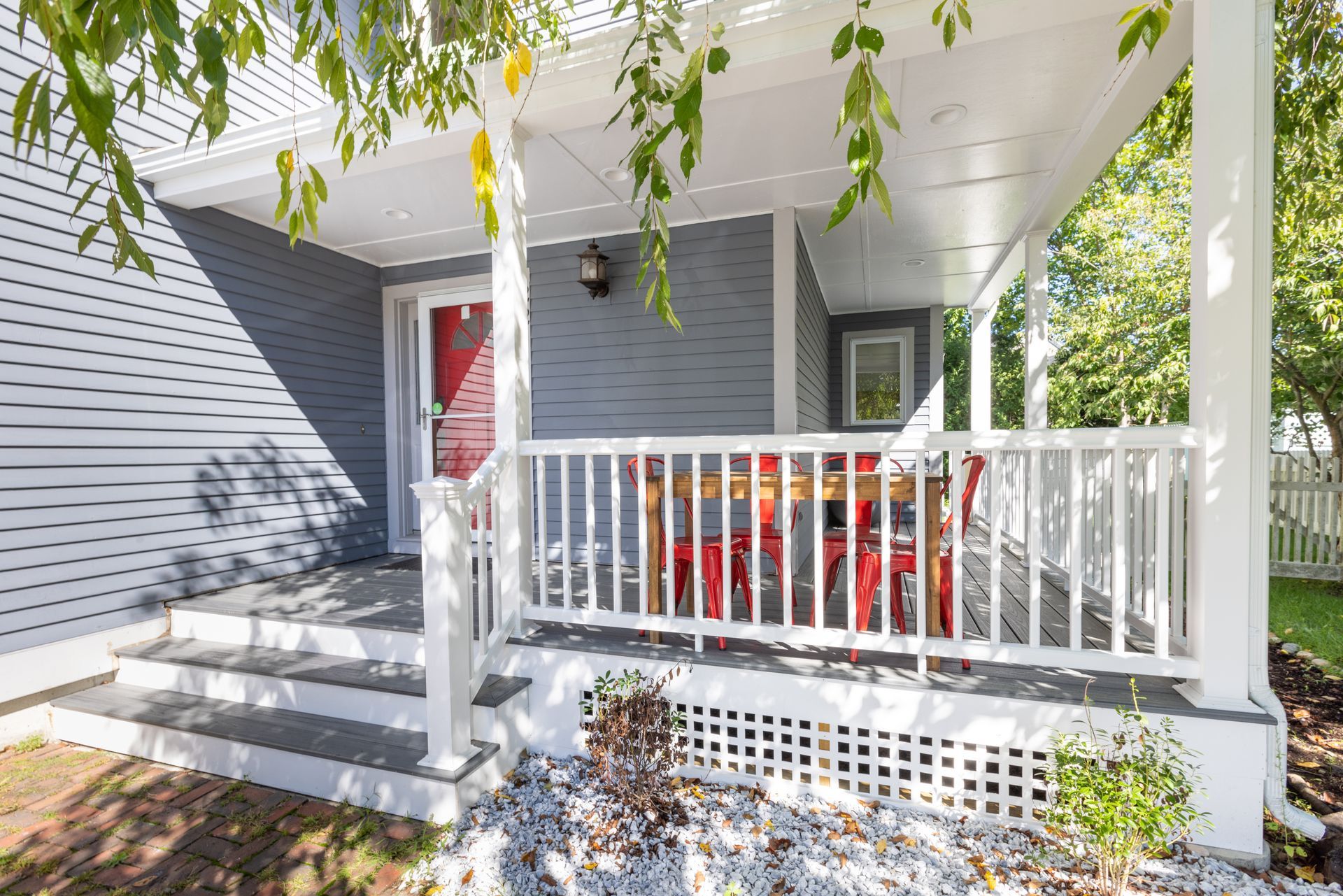 A blue house exterior featuring a wooden porch with red chairs, white railings, and steps leading to a red front door.
