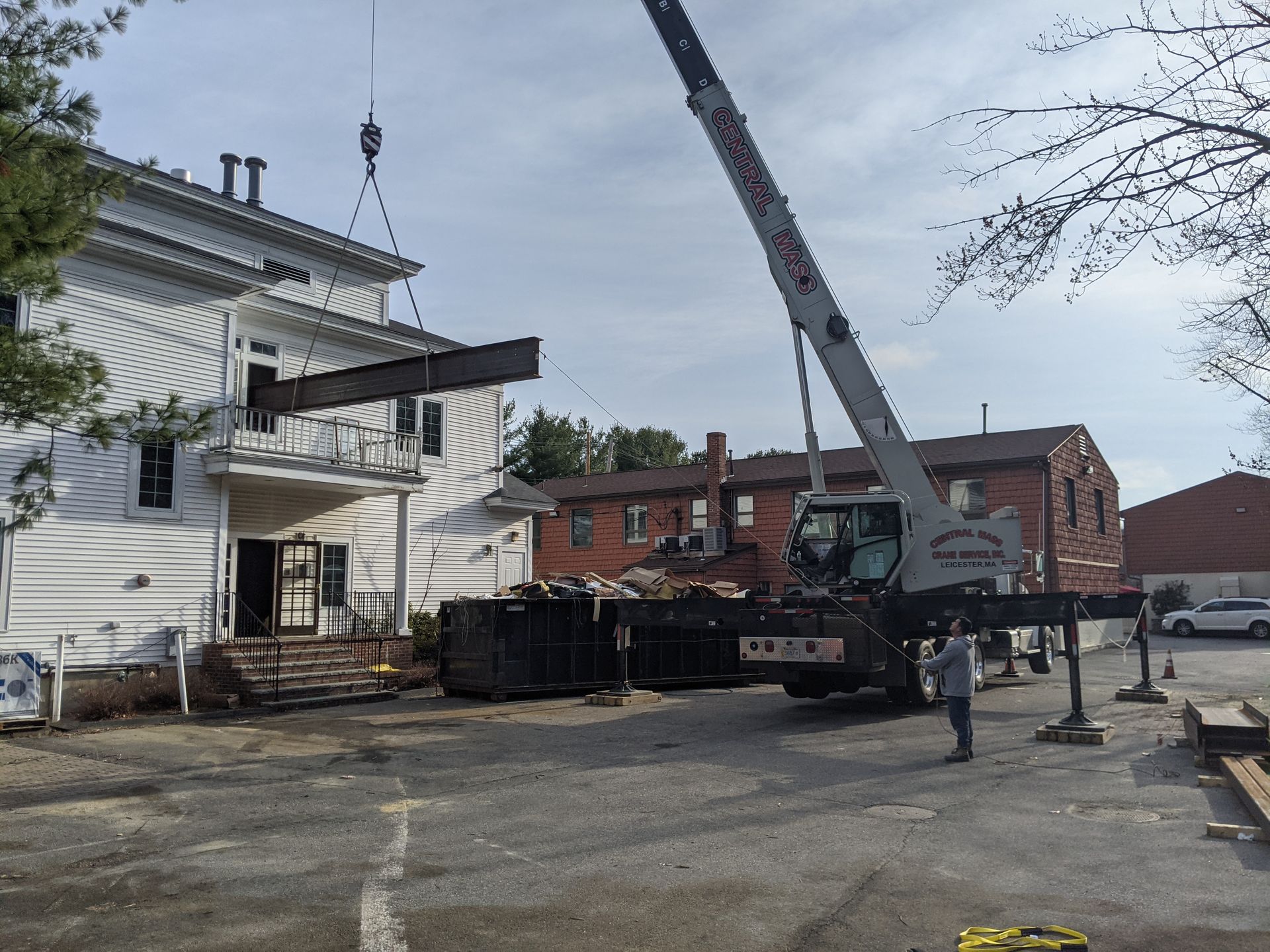 A crane lifts a large steel beam toward the porch of a white two-story building in a gravel lot.