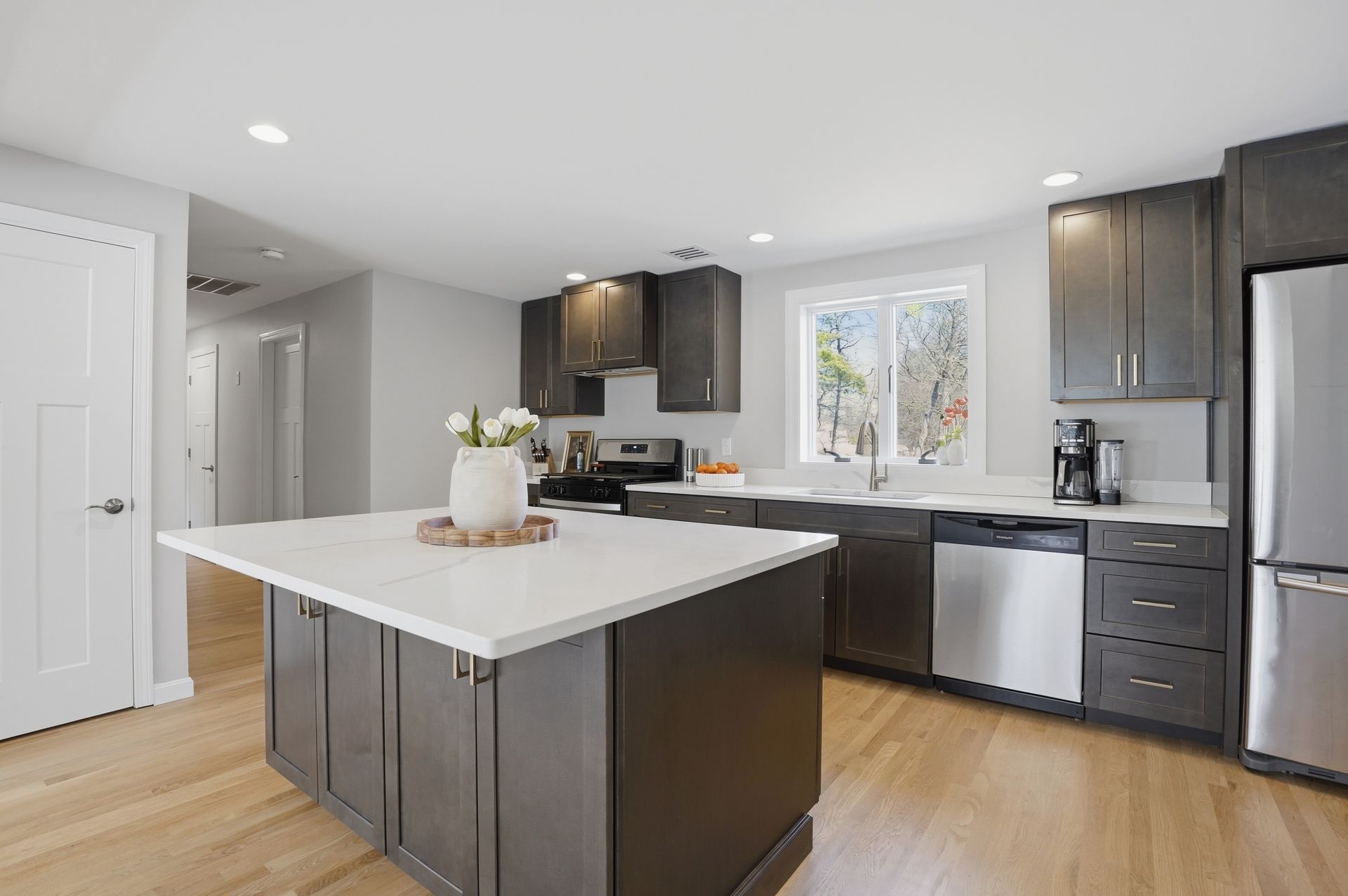 Bright modern kitchen with white island, dark cabinets, stainless appliances, and wood floors