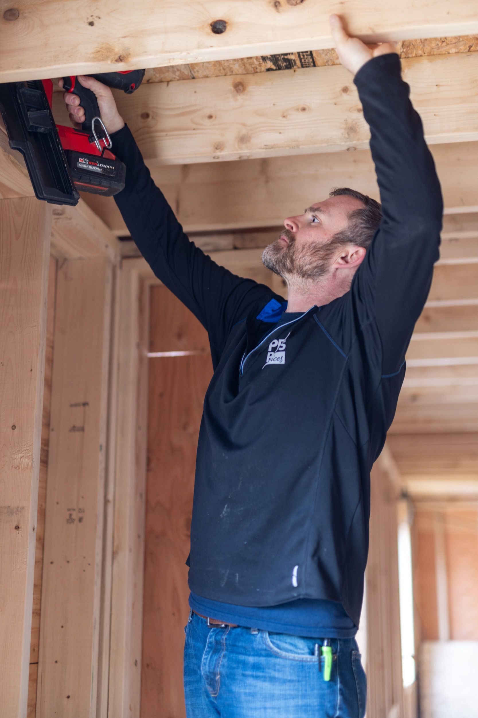 A person in a black long-sleeved shirt uses a power nailer to secure wooden framing on a construction site.