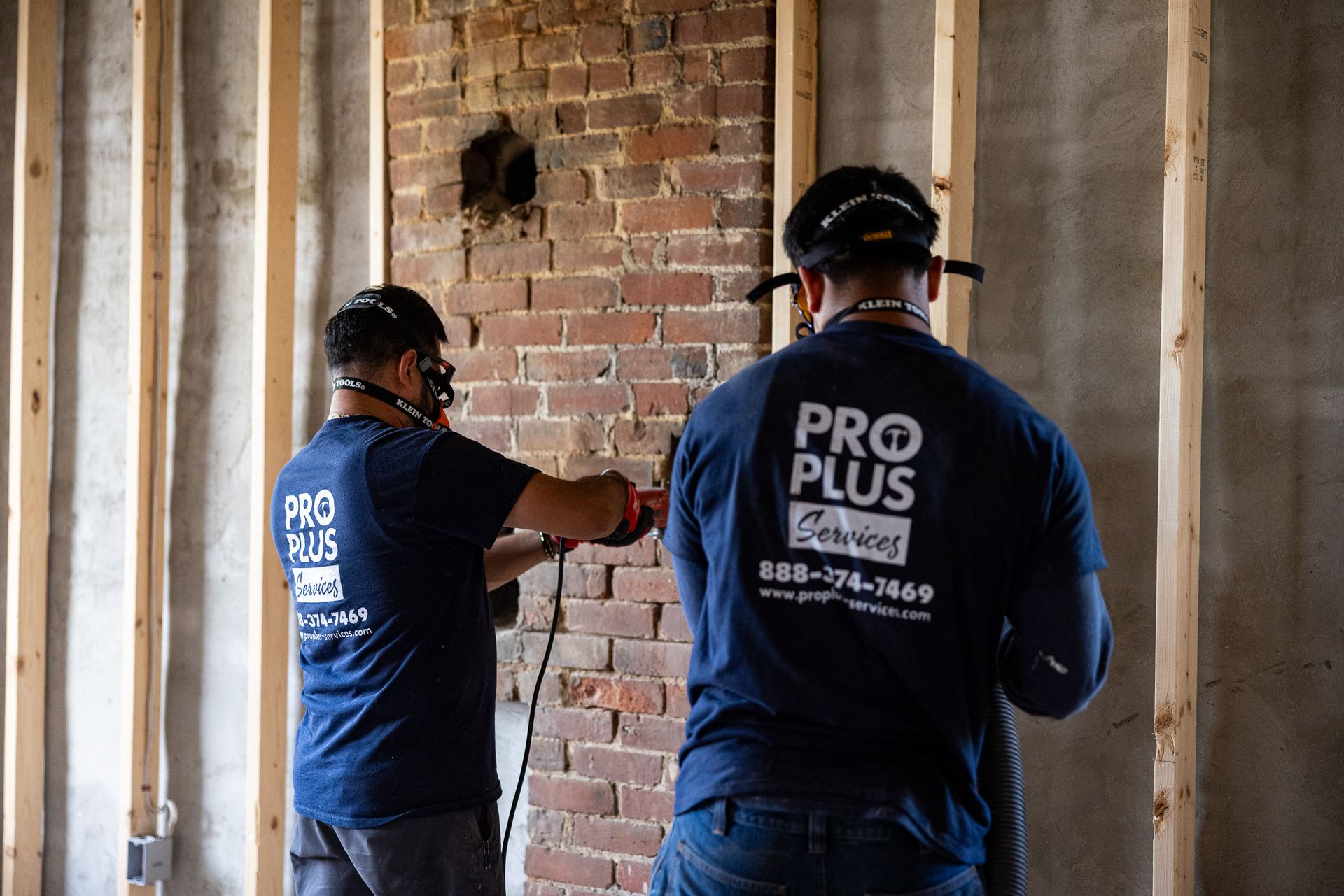 Two workers in navy Pro Plus Services shirts use tools on a brick chimney inside a room with unfinished wooden studs.